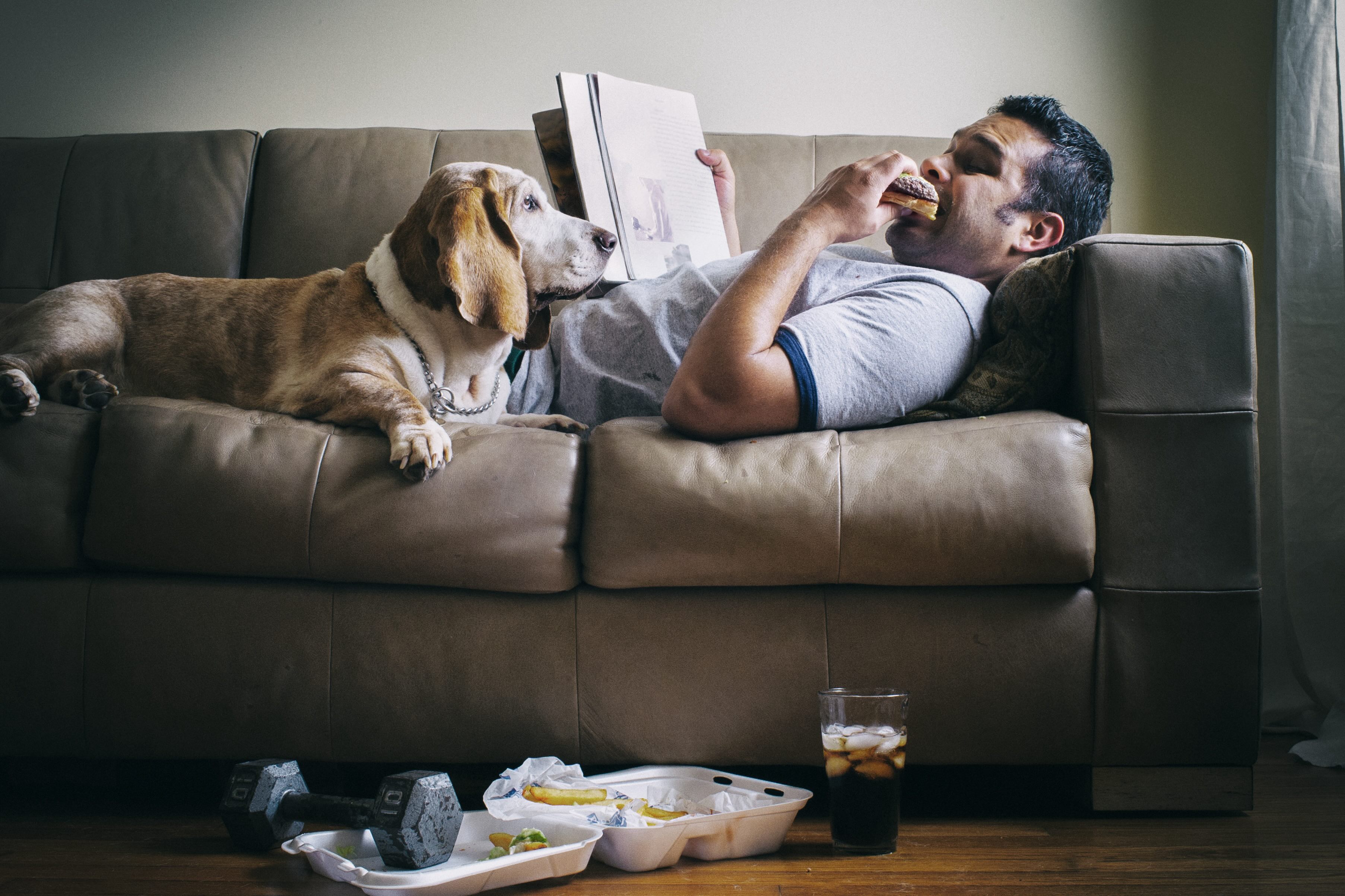A man eating a hamburger reading a fitness magazine while his dog watches.