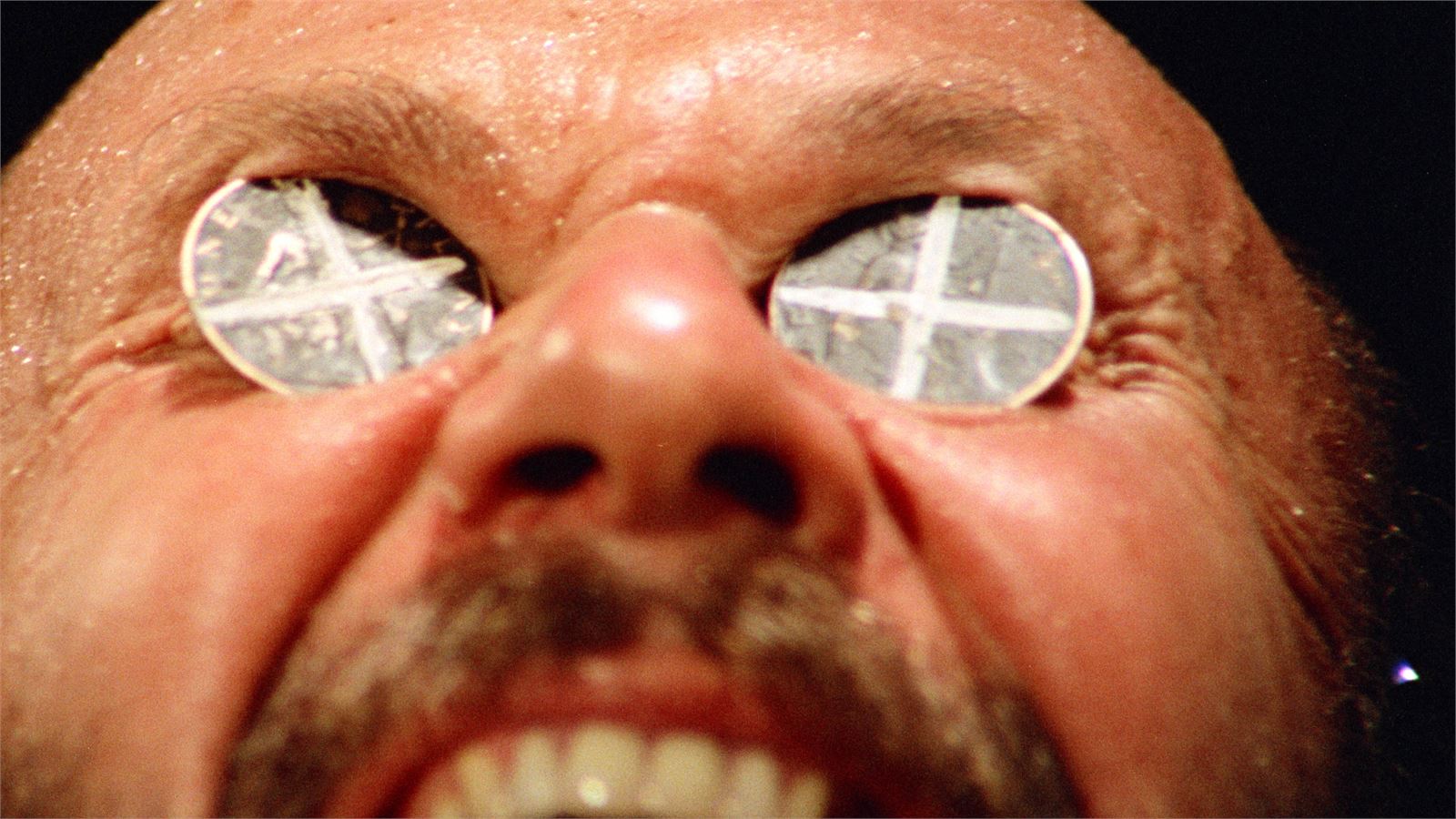 A movie still close-up of a man with two coins covering his eyes