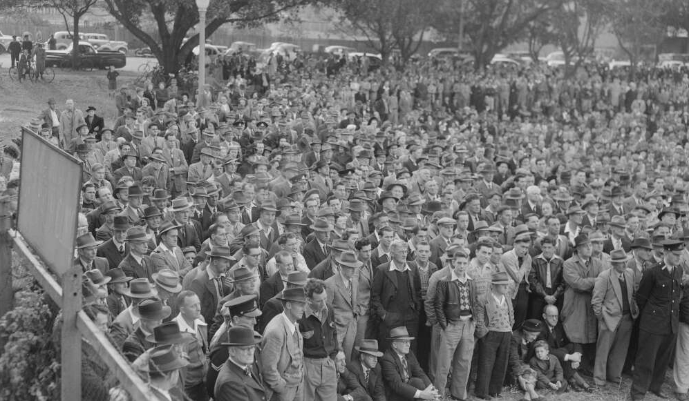 A black-and-white photo of a large group of men listening to a speaker