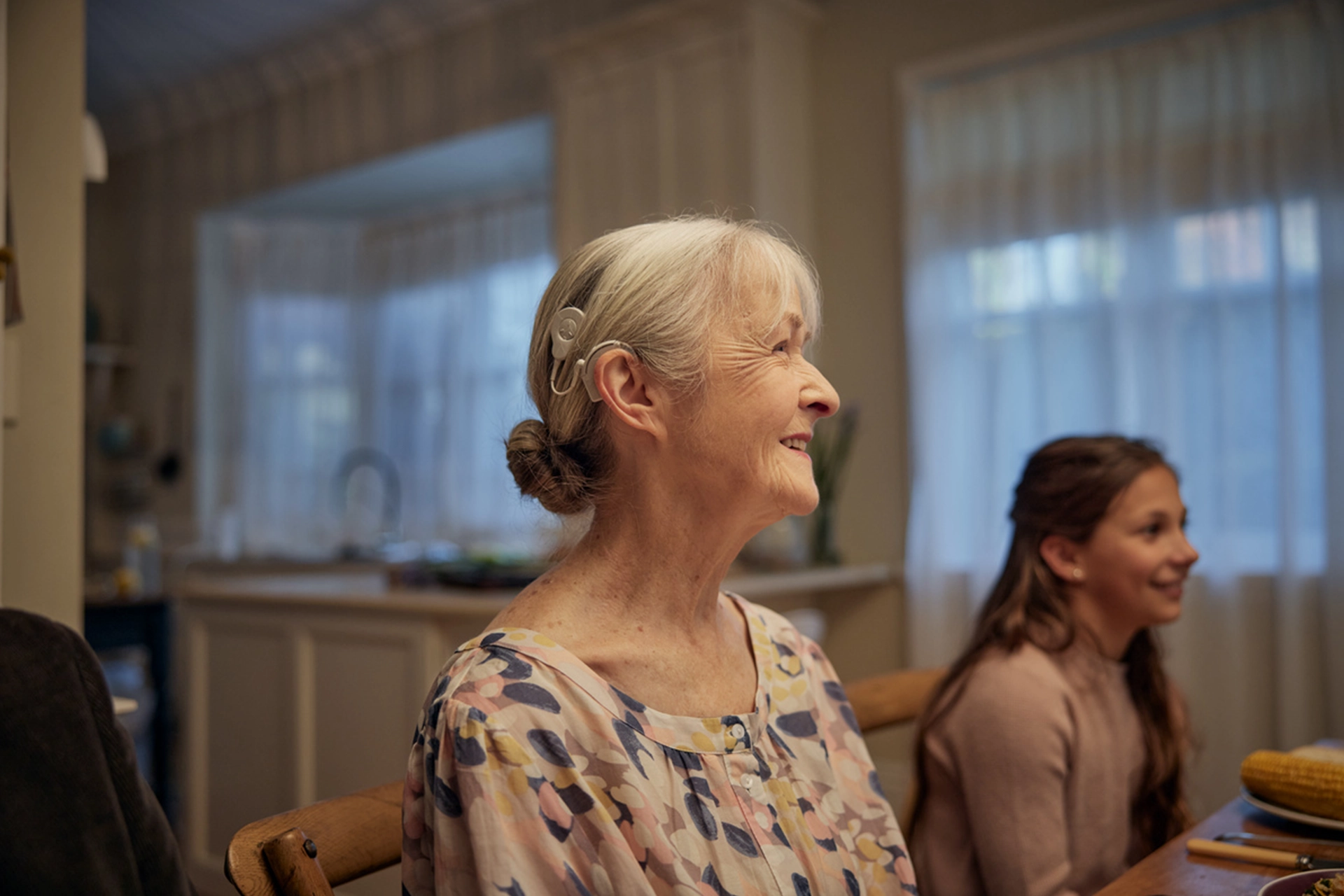 Elderly woman smiling and wearing a cochlear implant, sitting at dining table with granddaughter beside her