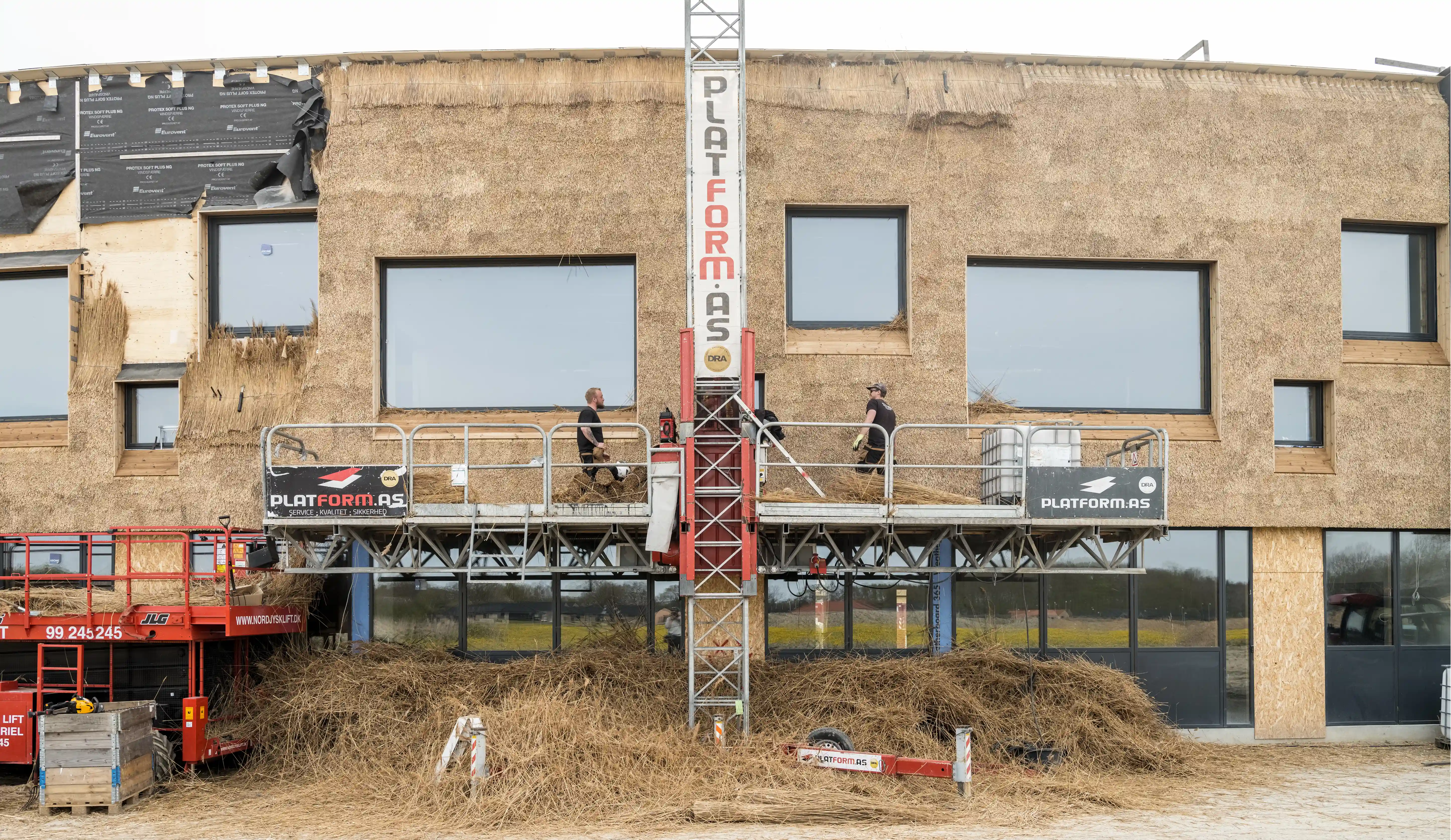 A school being built with tightly bound straw on the outside walls. Two men are working from a raised platform. A large stack of loose hay sits below their platform.