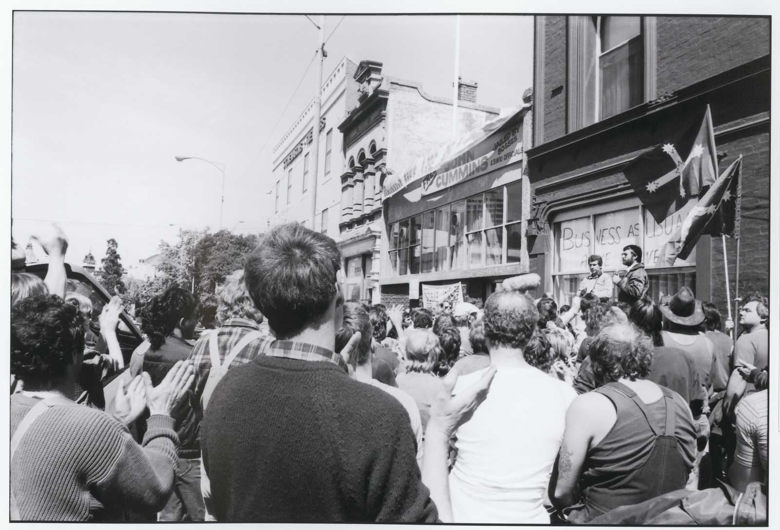 A black-and-white photo of a crowd listening to a speaker in the street