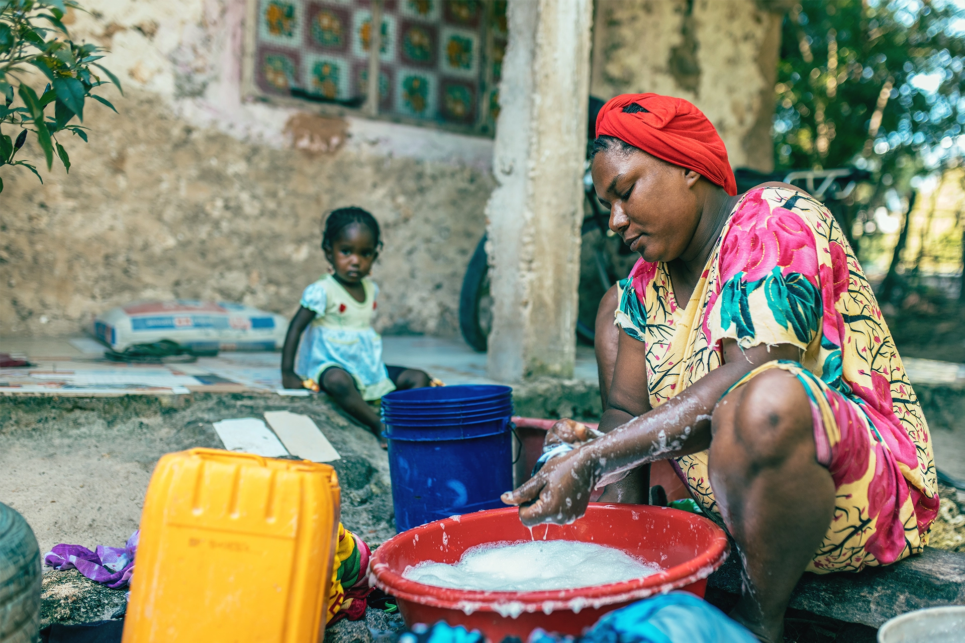 African woman sitting washing clothes in a bucket with young girl in background