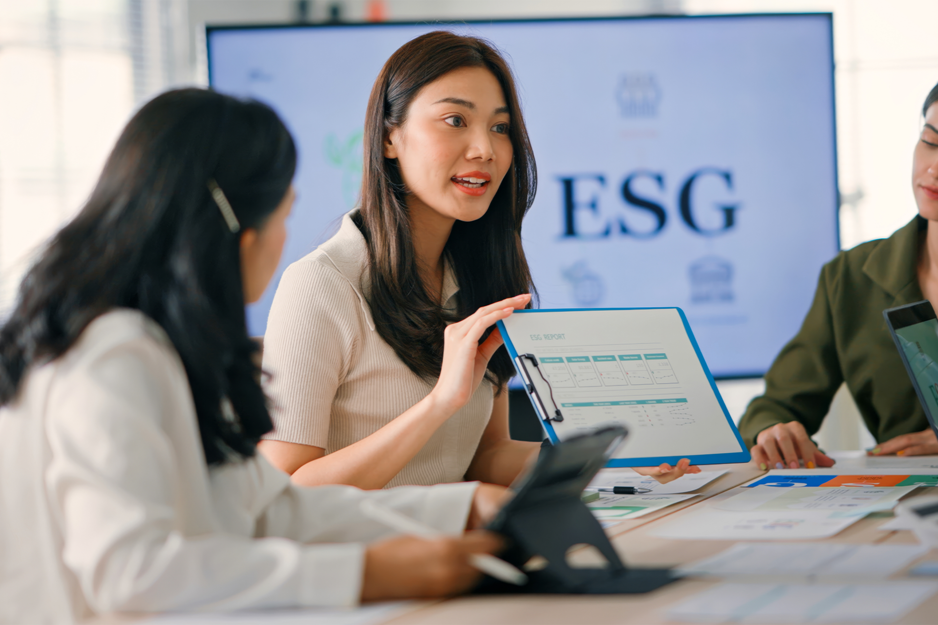 A woman presenting to two other women around an office table, with a television with ESG written on it in the background