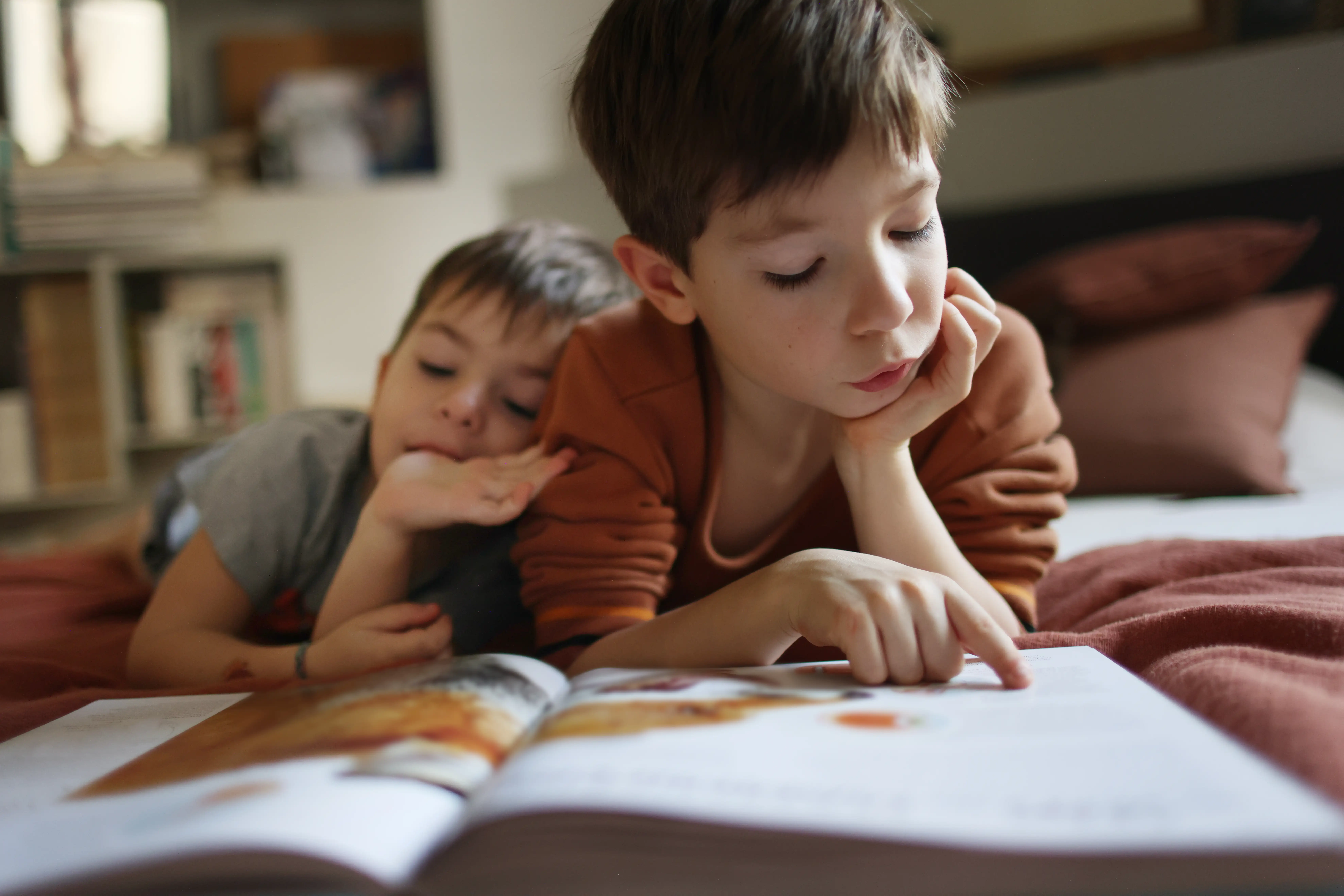 Two young brothers lying on a bed reading a book together