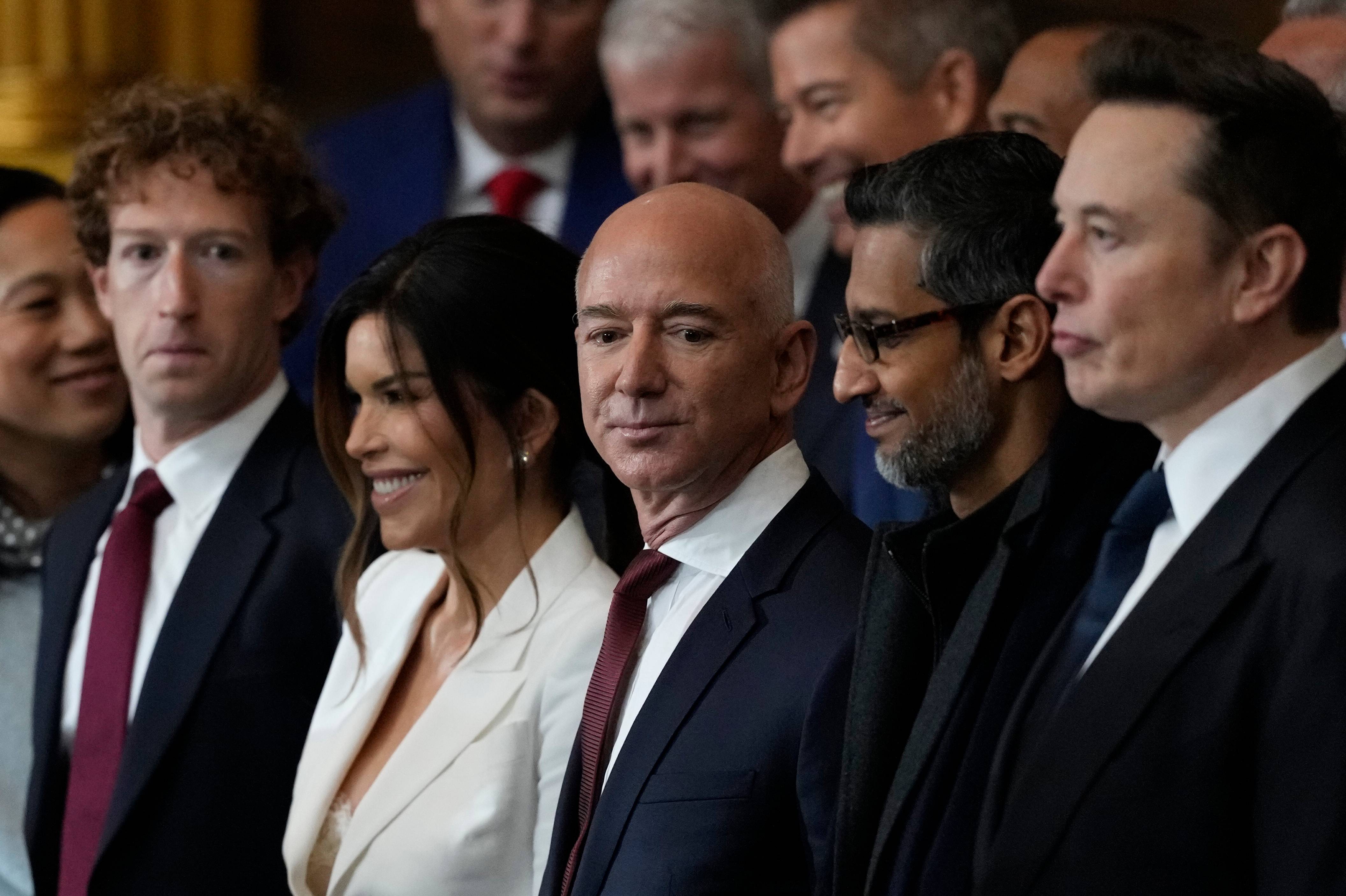 American billionaires Mark Zuckerberg, Lauren Sanchez, Jeff Bezos, Sundar Pichai and Elon Musk stand in a line in suits. 