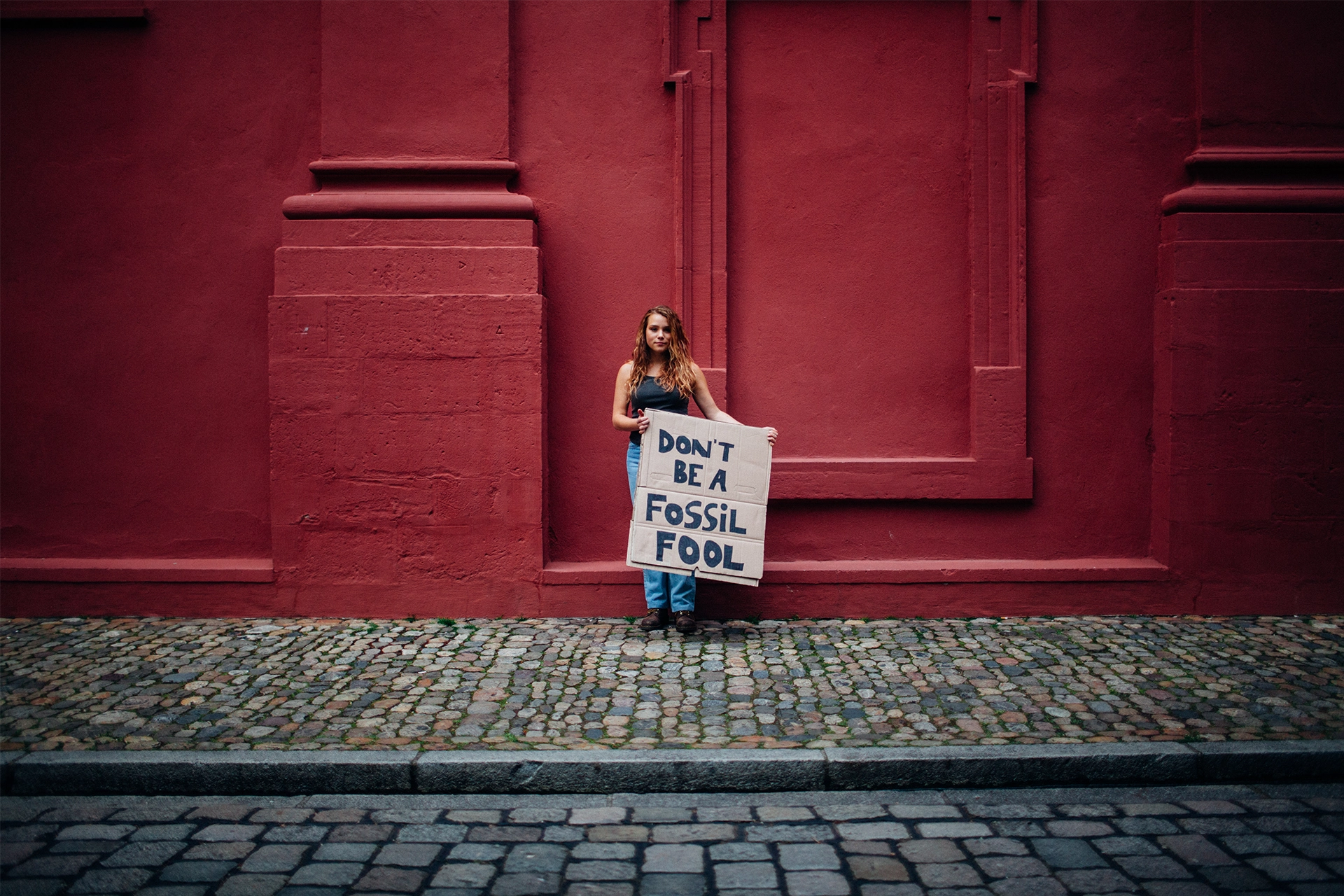 Woman standing against red painted wall holding sign that says: Don't be a fossil fool