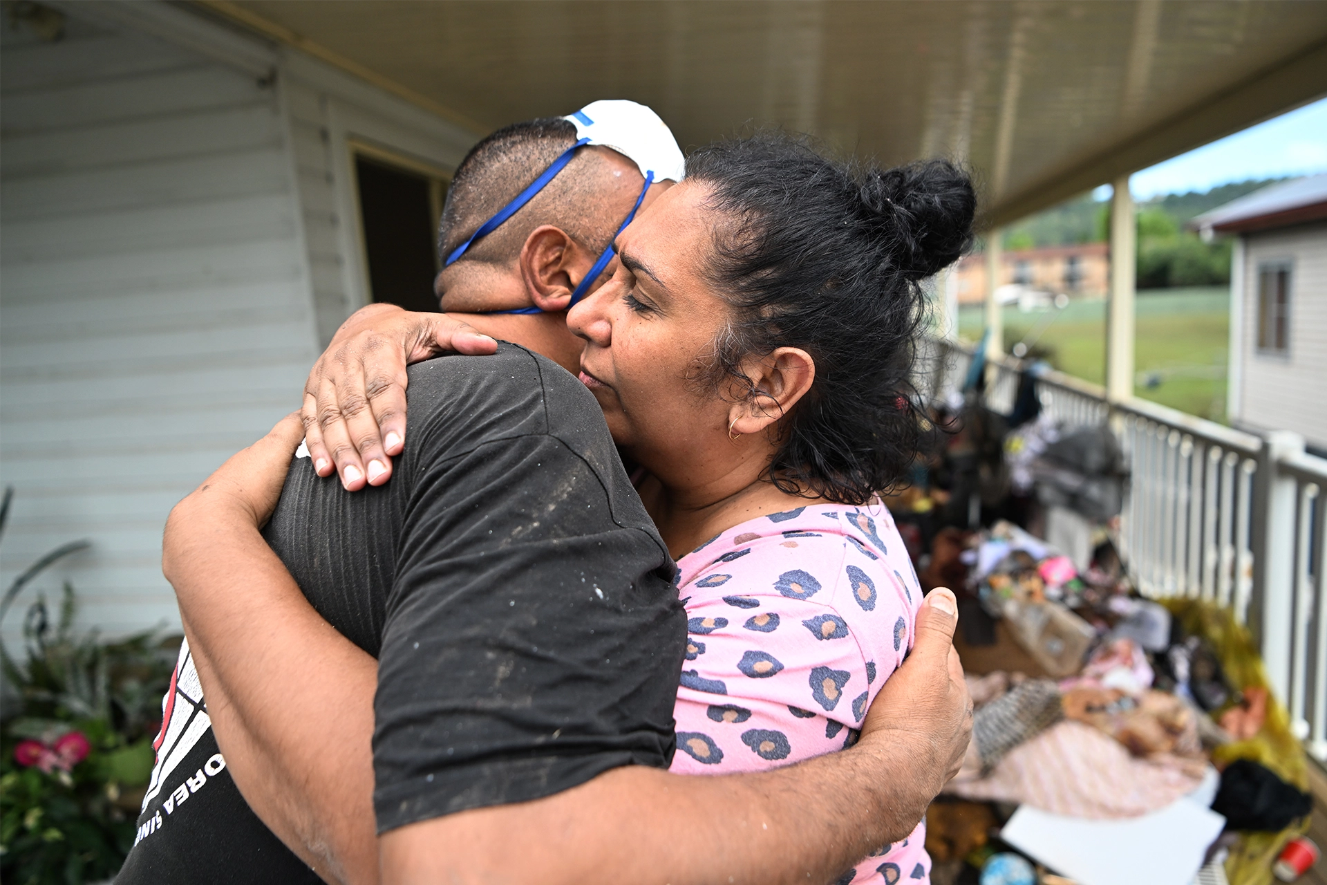 Man and woman hugging in front of a recently flooded house, with possessions on the verandah outside