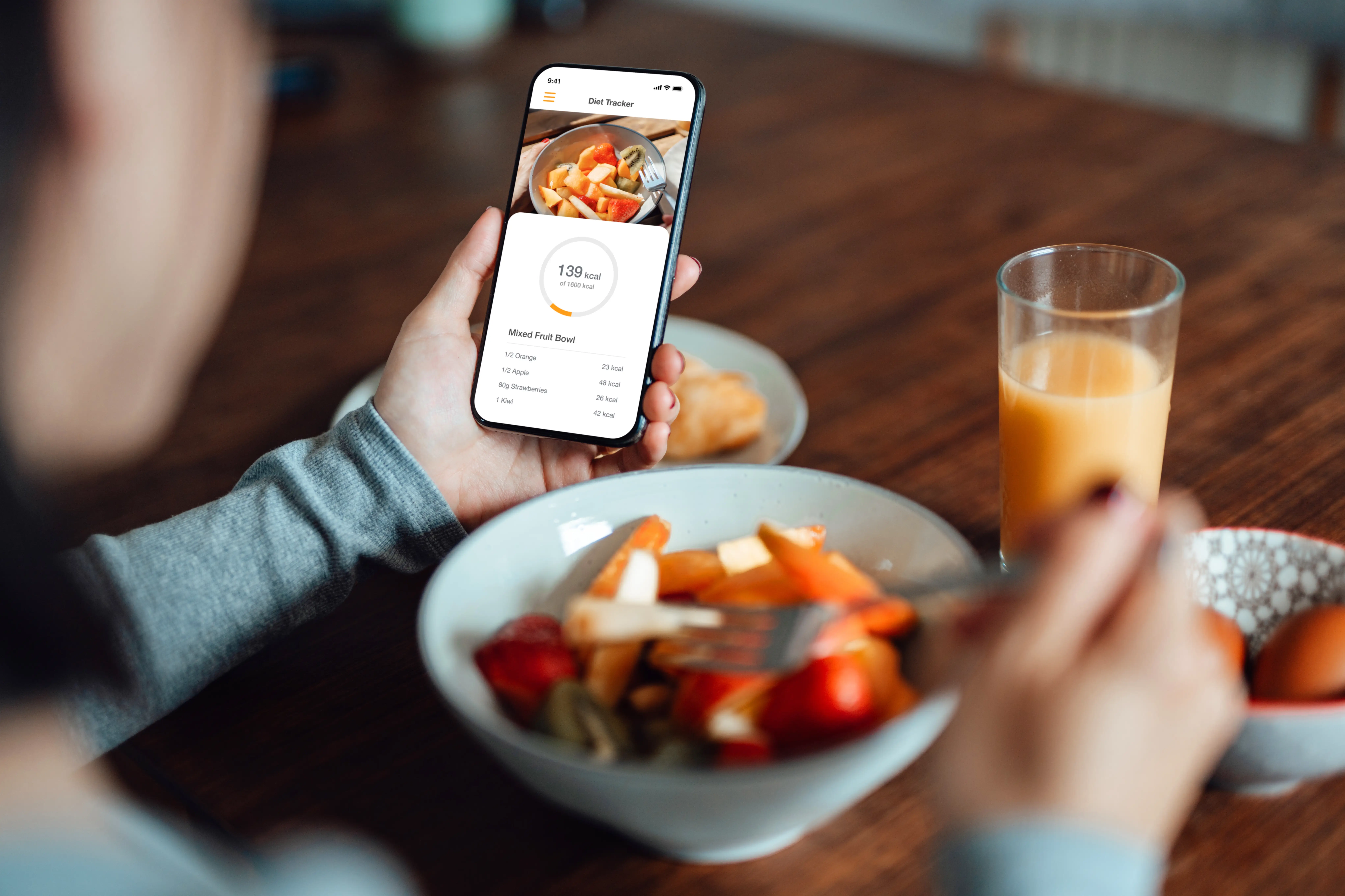 Over the shoulder shot of young woman using mobile app to track nutrition and count calories with smartphone while eating a healthy breakfast of fruit
