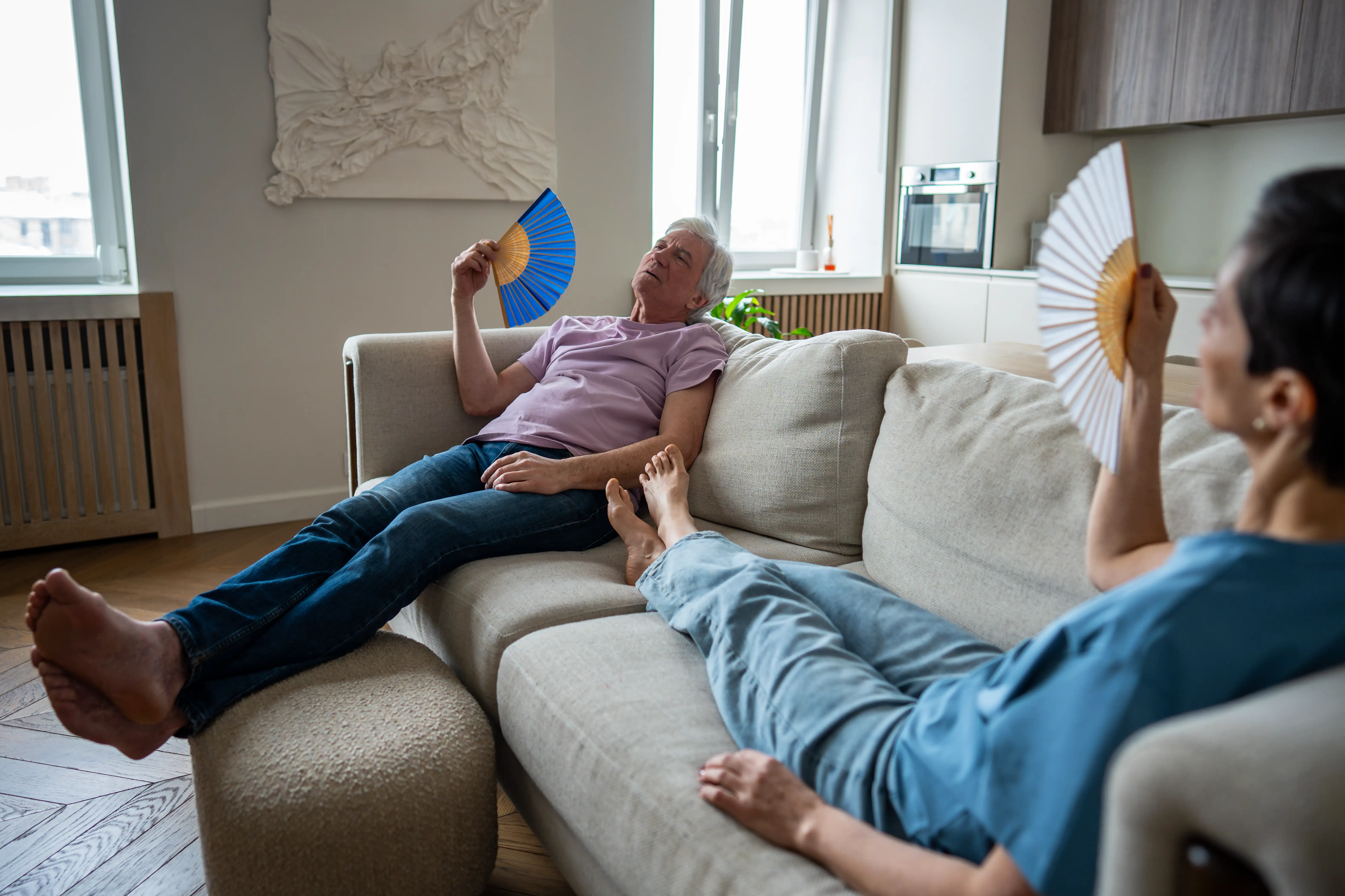 Tired senior couple in extreme heat, using fans while lounging on couch