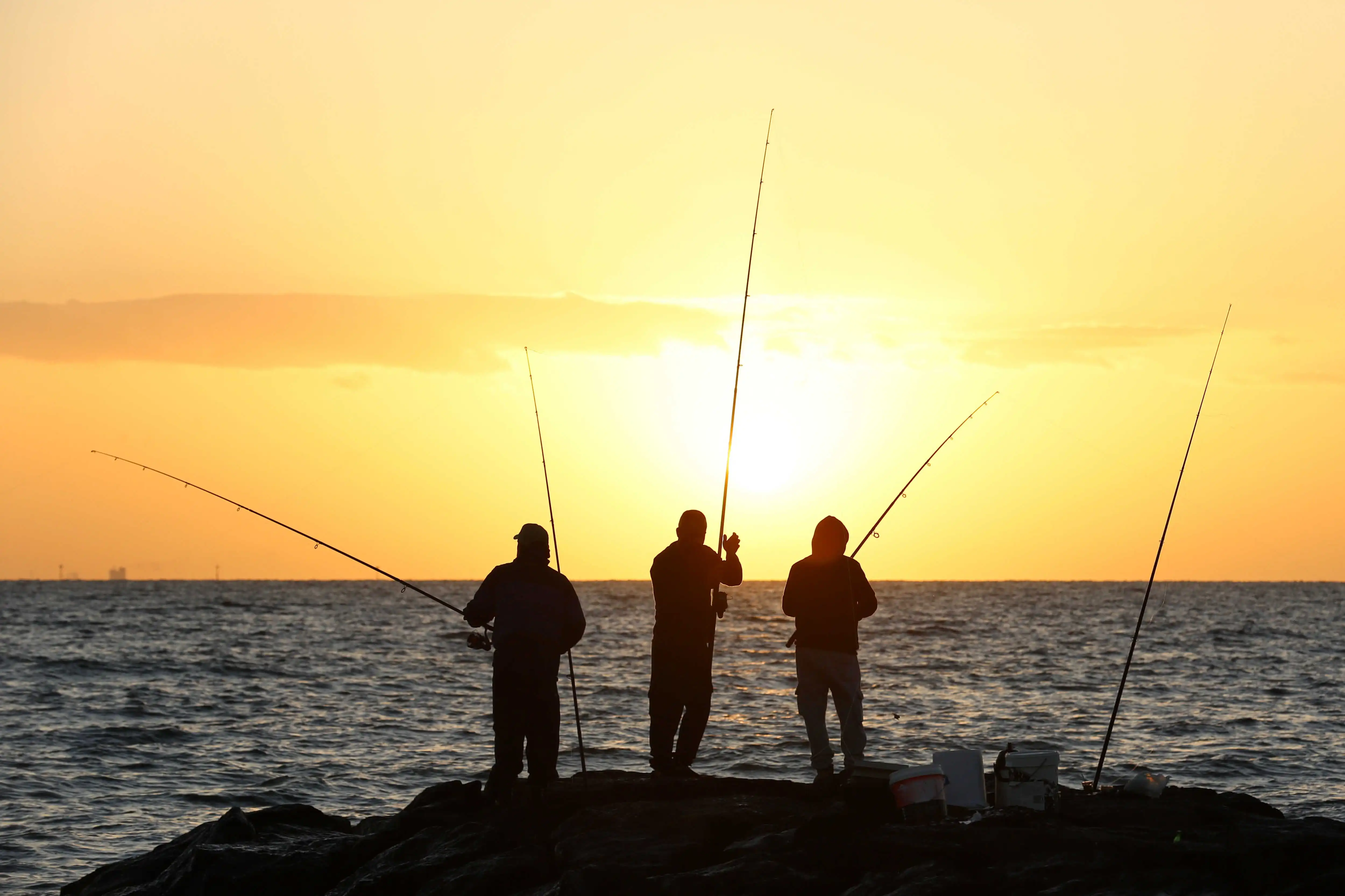 Silhouettes of three men fishing off rocks at sunset