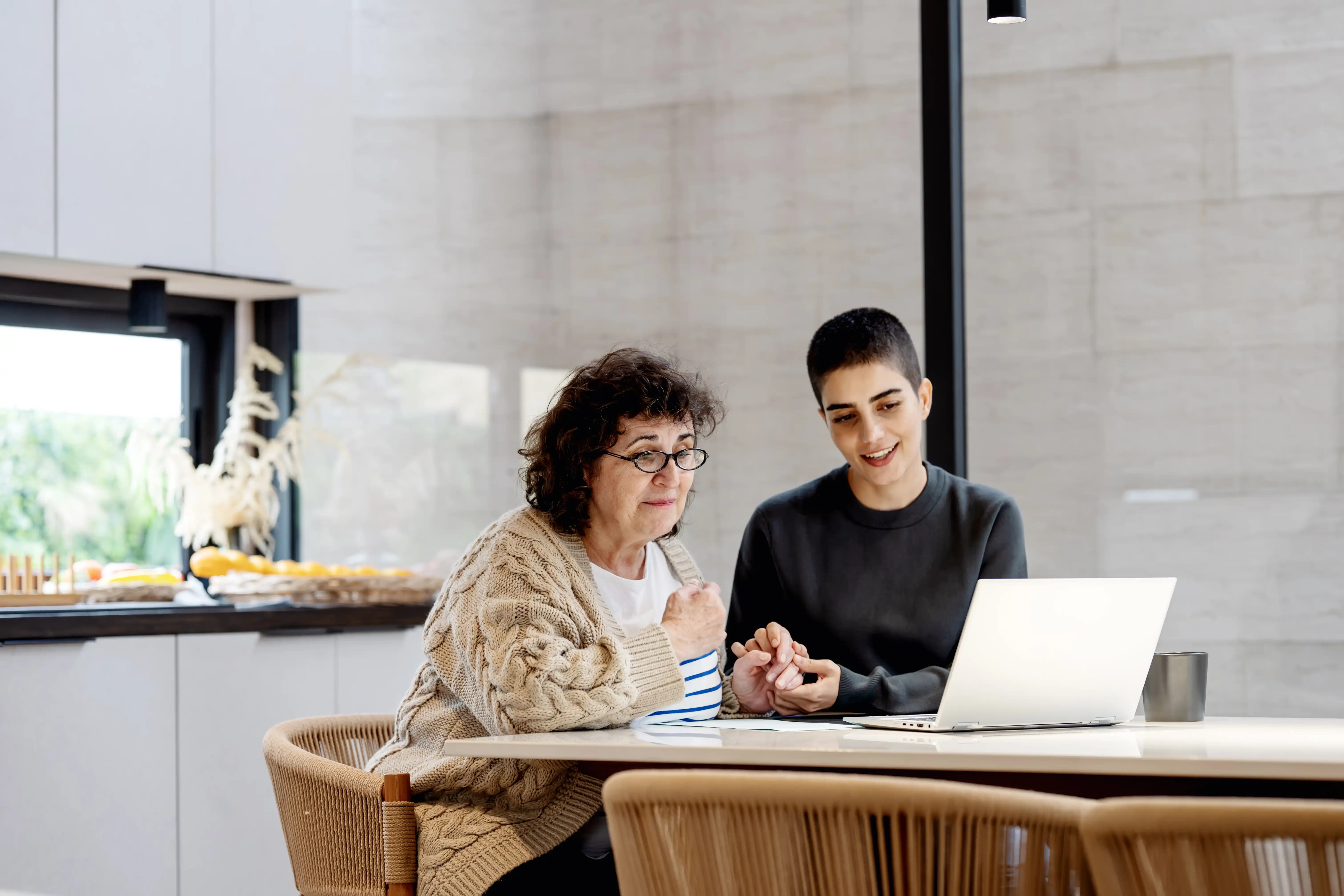 A daughter holds her mother's hand sitting at a dining table facing a laptop