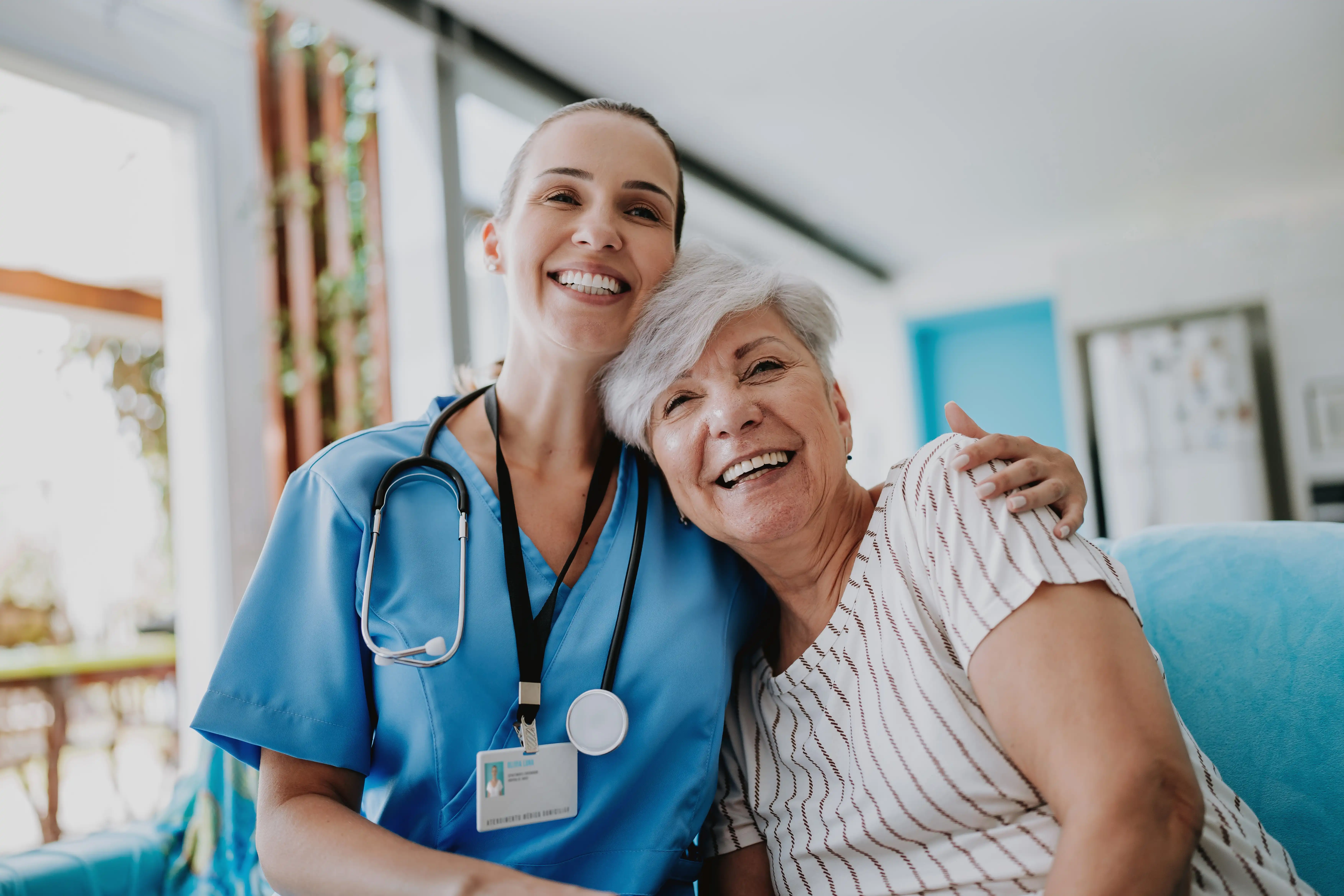 A female healthcare professional in scrubs hugs an older woman smiling arm in arm