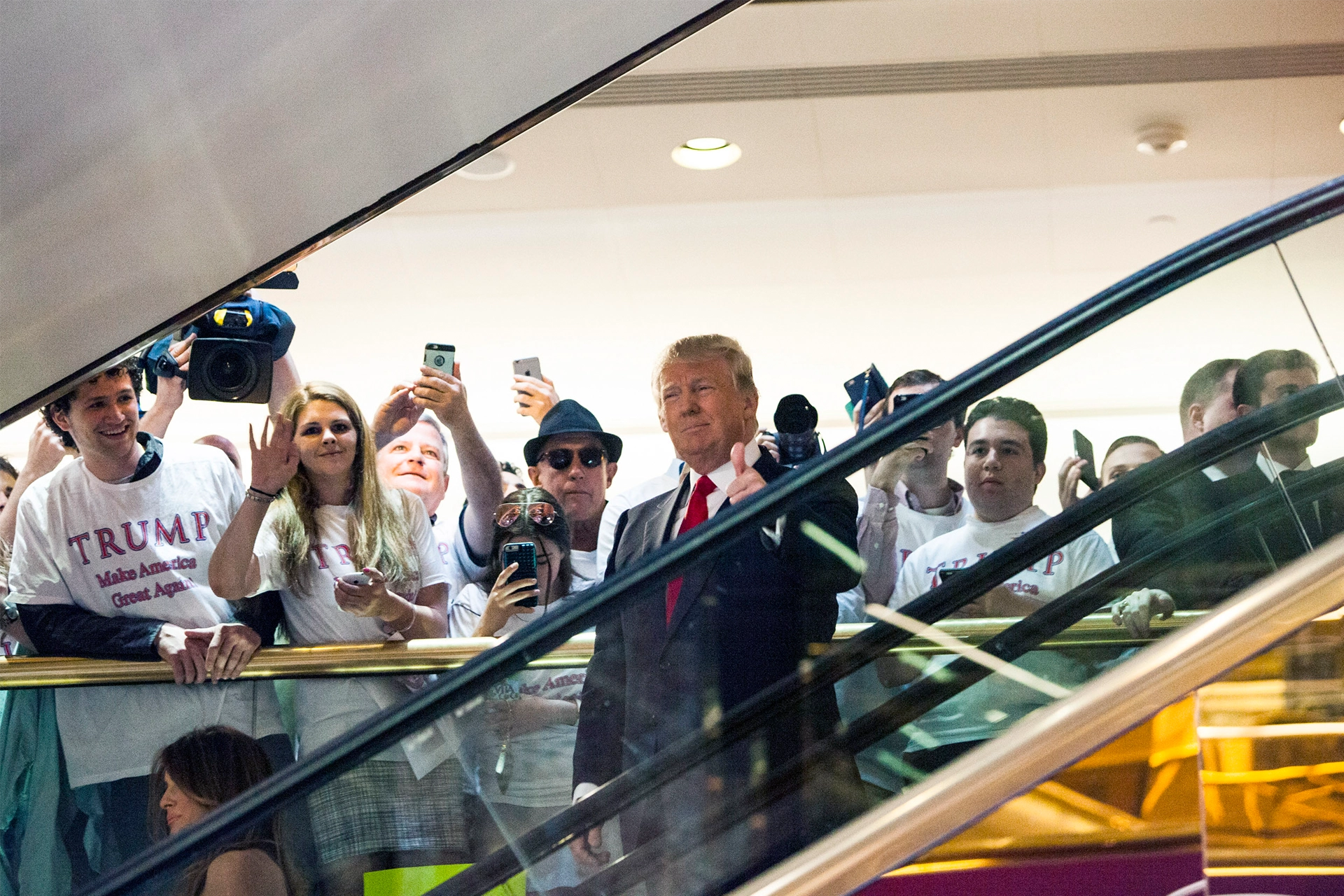 Donald Trump descending an escalator giving the thumbs up, with crowd of supports in background