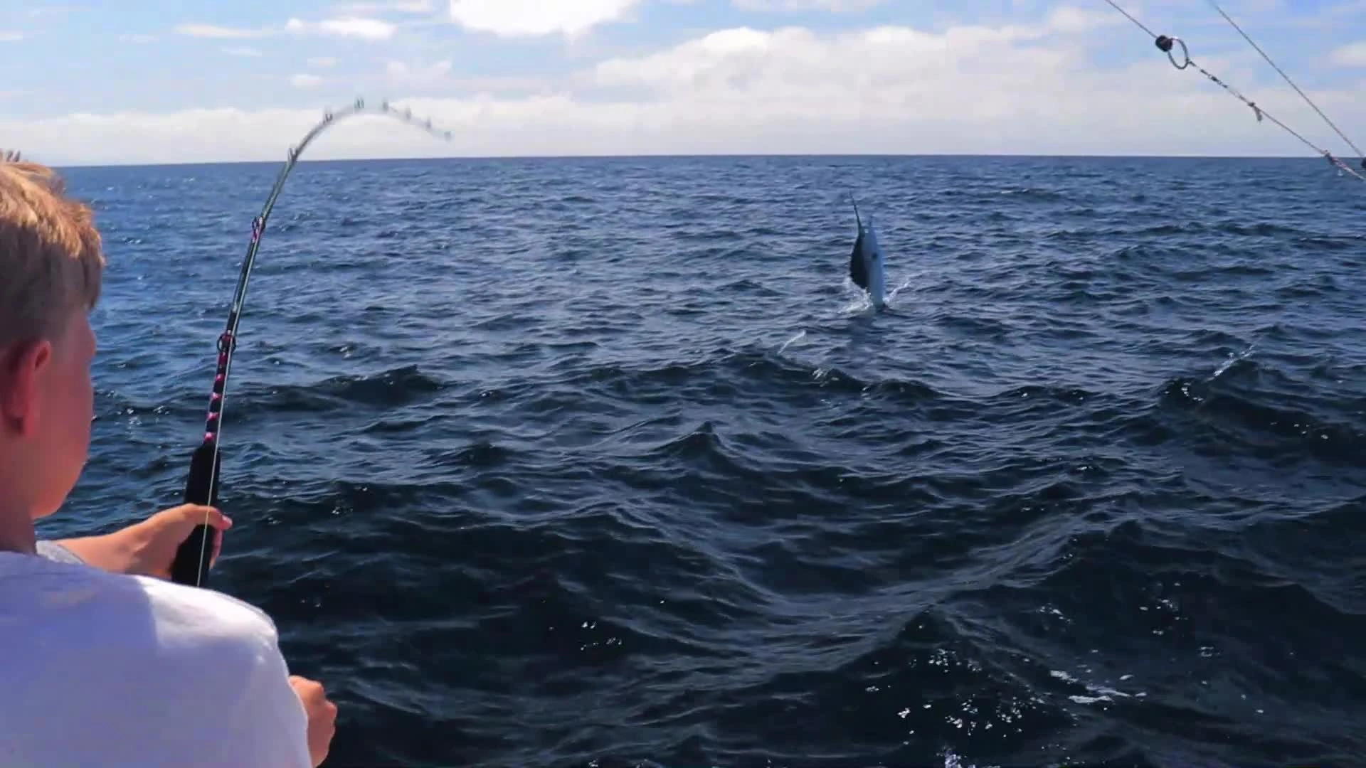A boy reels in a large fish off the back of a boat which is bursting out of the water