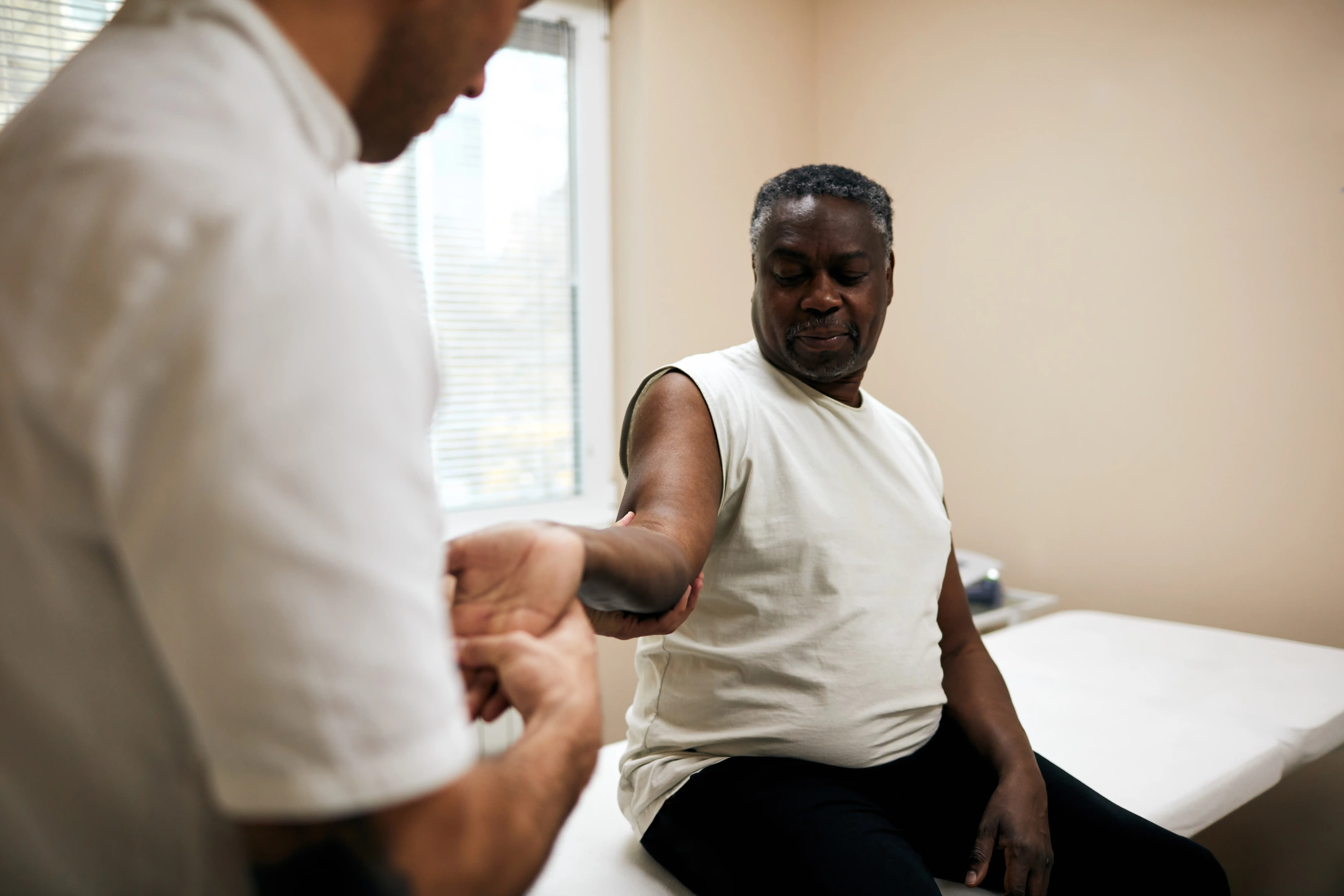 A man sitting on a bed in a medical clinic holds his arm out for the doctor to inspect