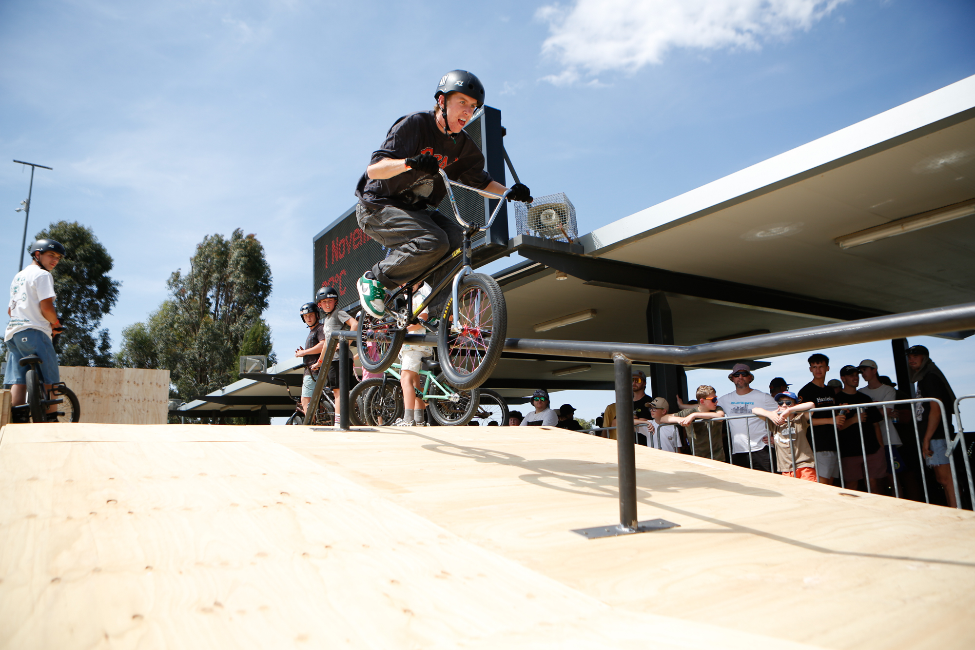 A young BMX rider grinds along a pole at a skate competition under sunny blue skies. He is holding onto the handlebars tightly and concentrating as people look on from behind a barrier