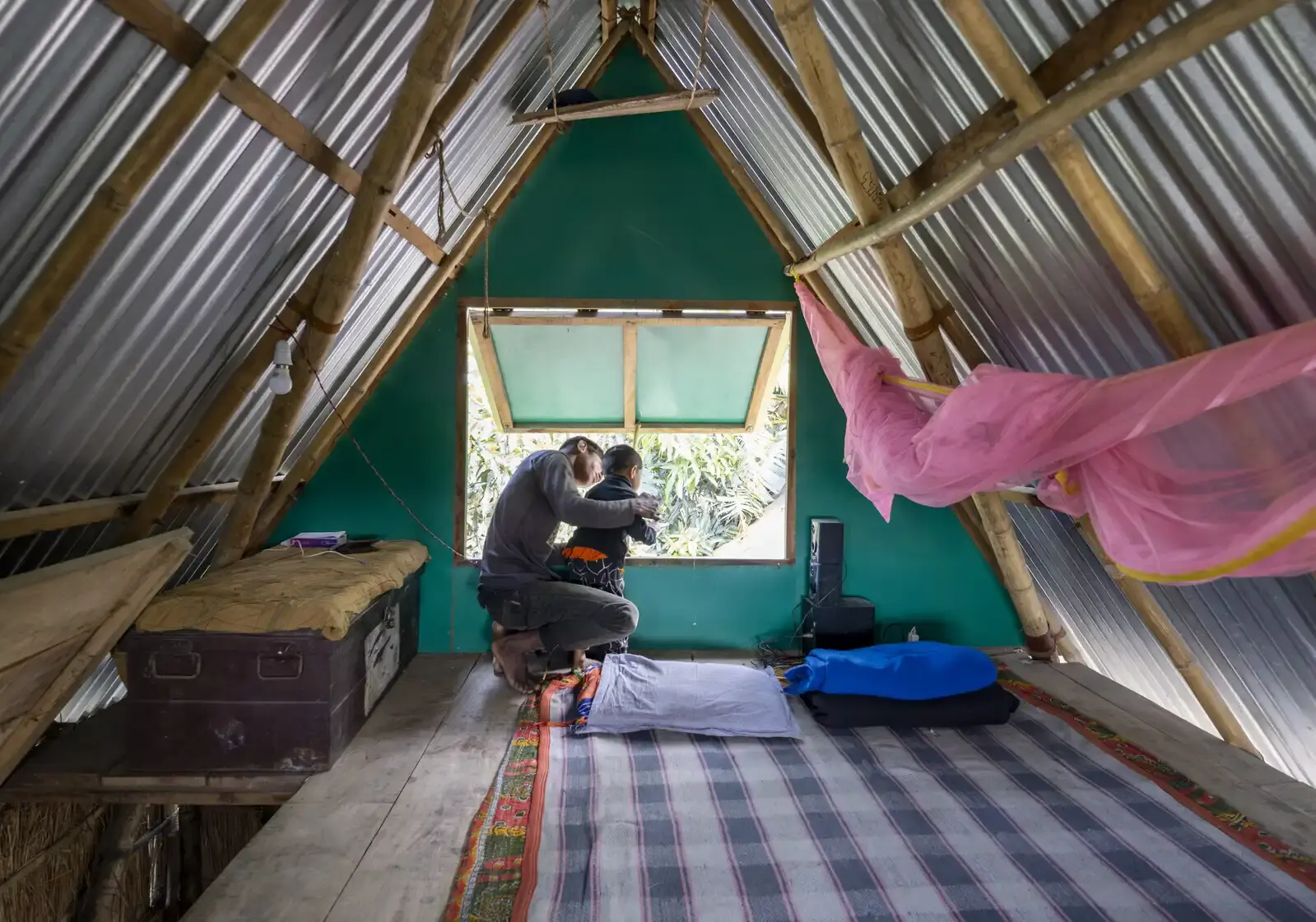 A father and young child look out the window of an elevated bamboo house. The house is made from bamboo and sheets of metal with a chest in the corner and blankets and pillows on the floor.