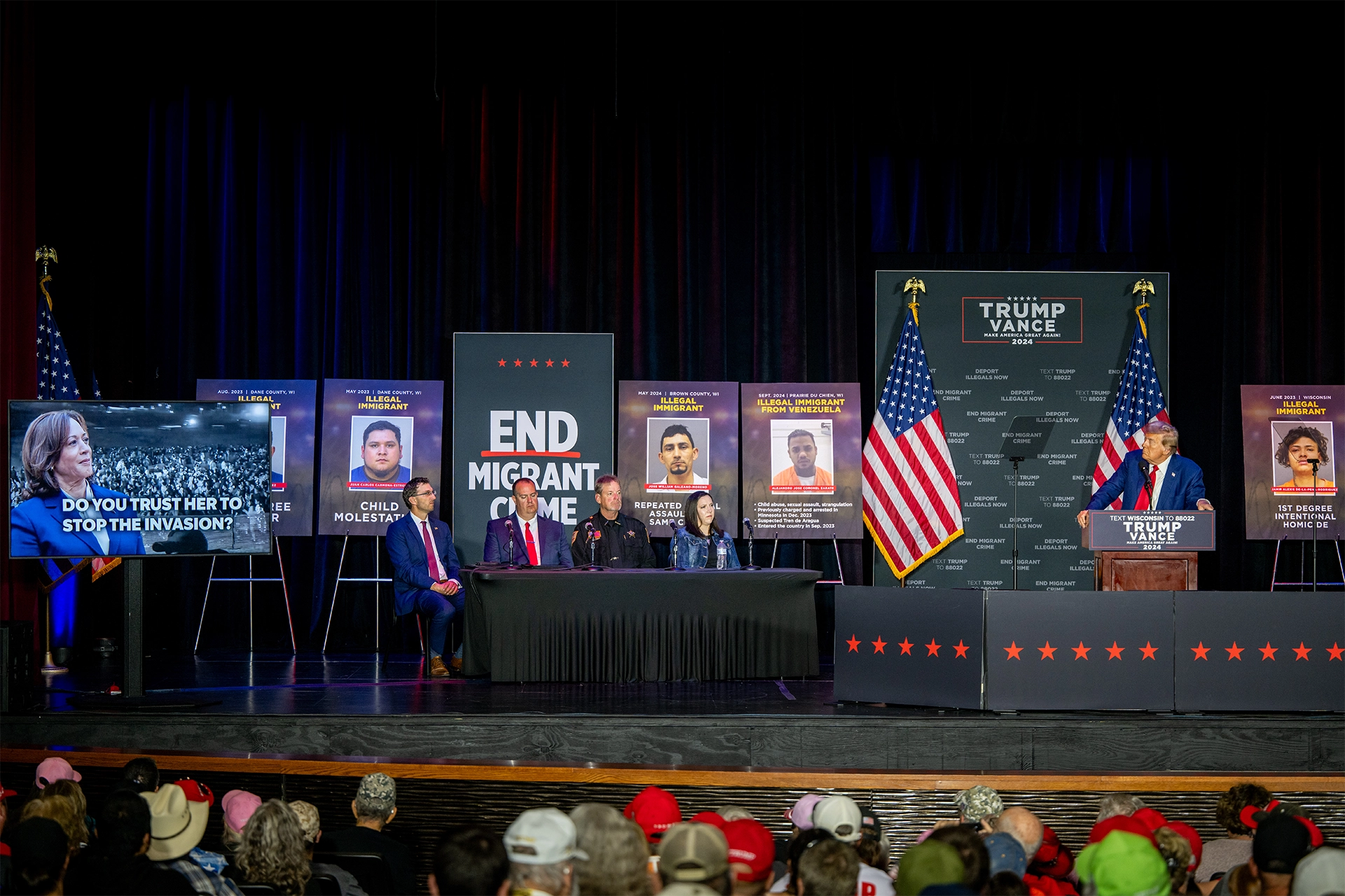 Donald Trump on stage delivering a speech, with signs about migrants in background