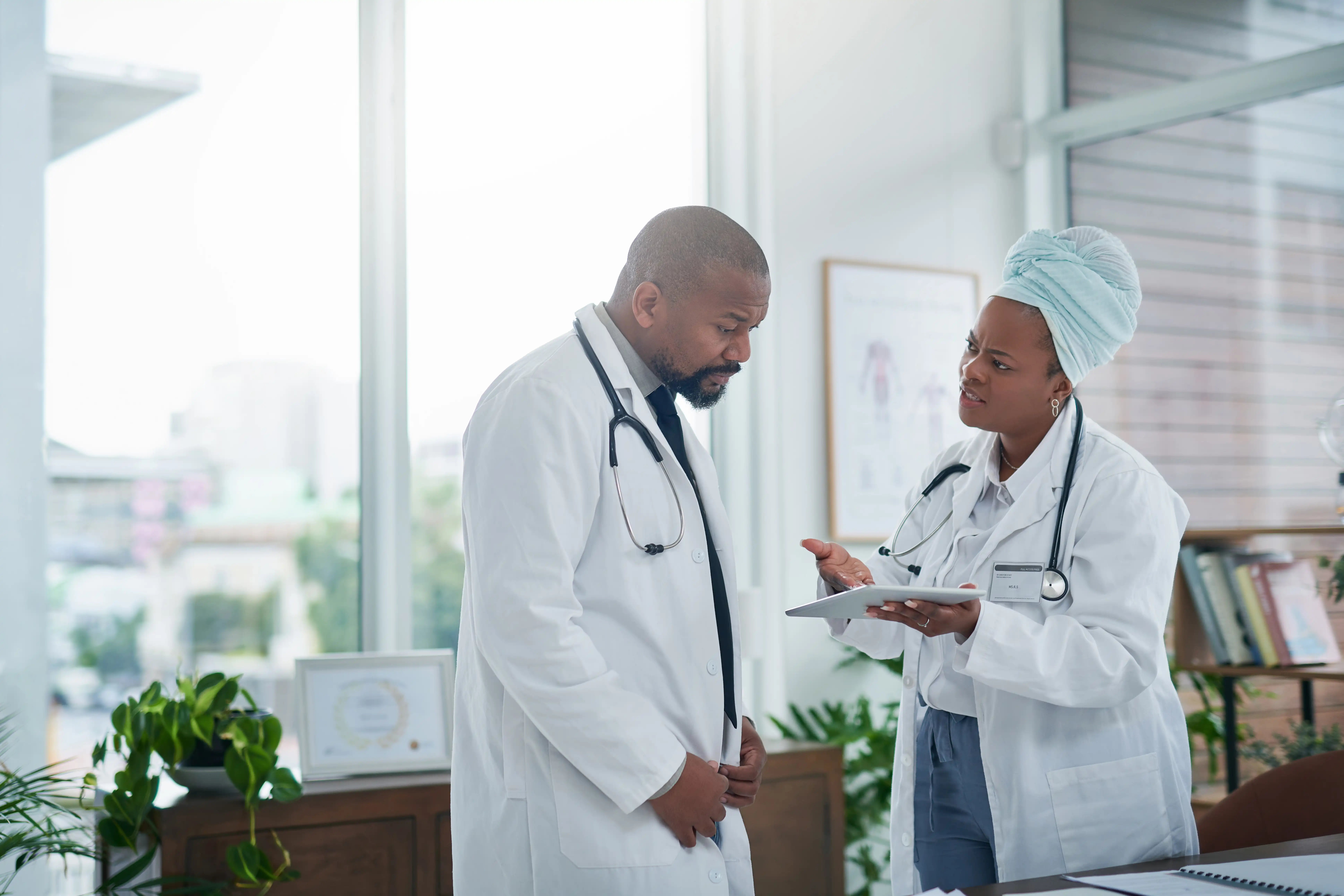 Two doctors at a hospital looking at a digital tablet device, and one of them is pointing at the screen with a confused expression