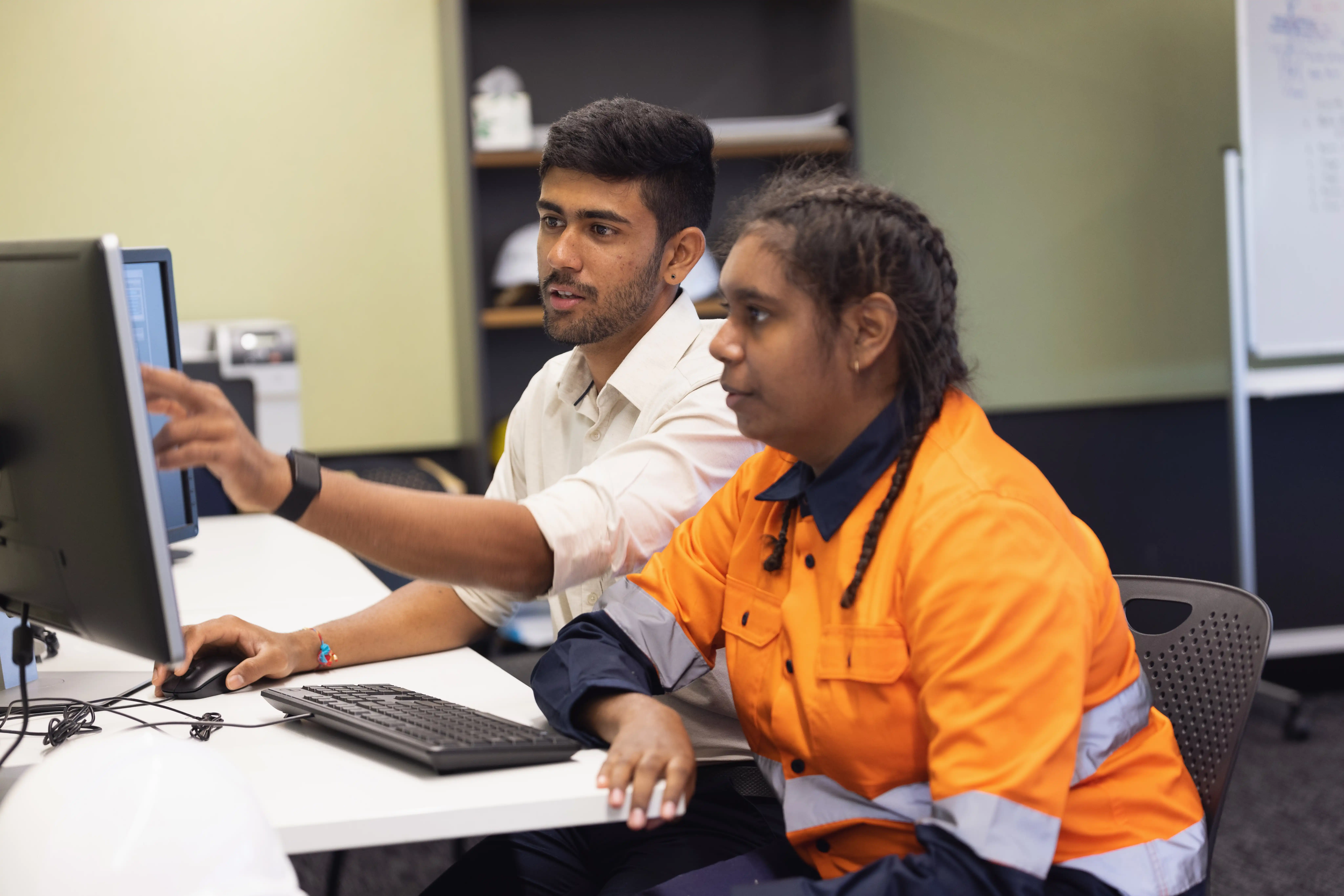 An Indian-Australian man and an Aboriginal Australian woman wearing high vis sitting at a desk looking at a computer screen