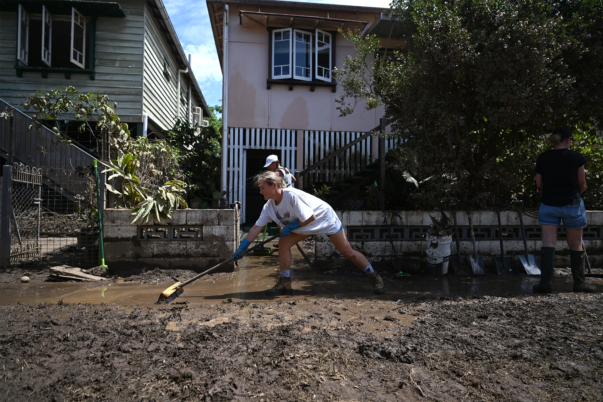 Woman wearing boots and gloves sweeping away mud in front of a recently flooded house