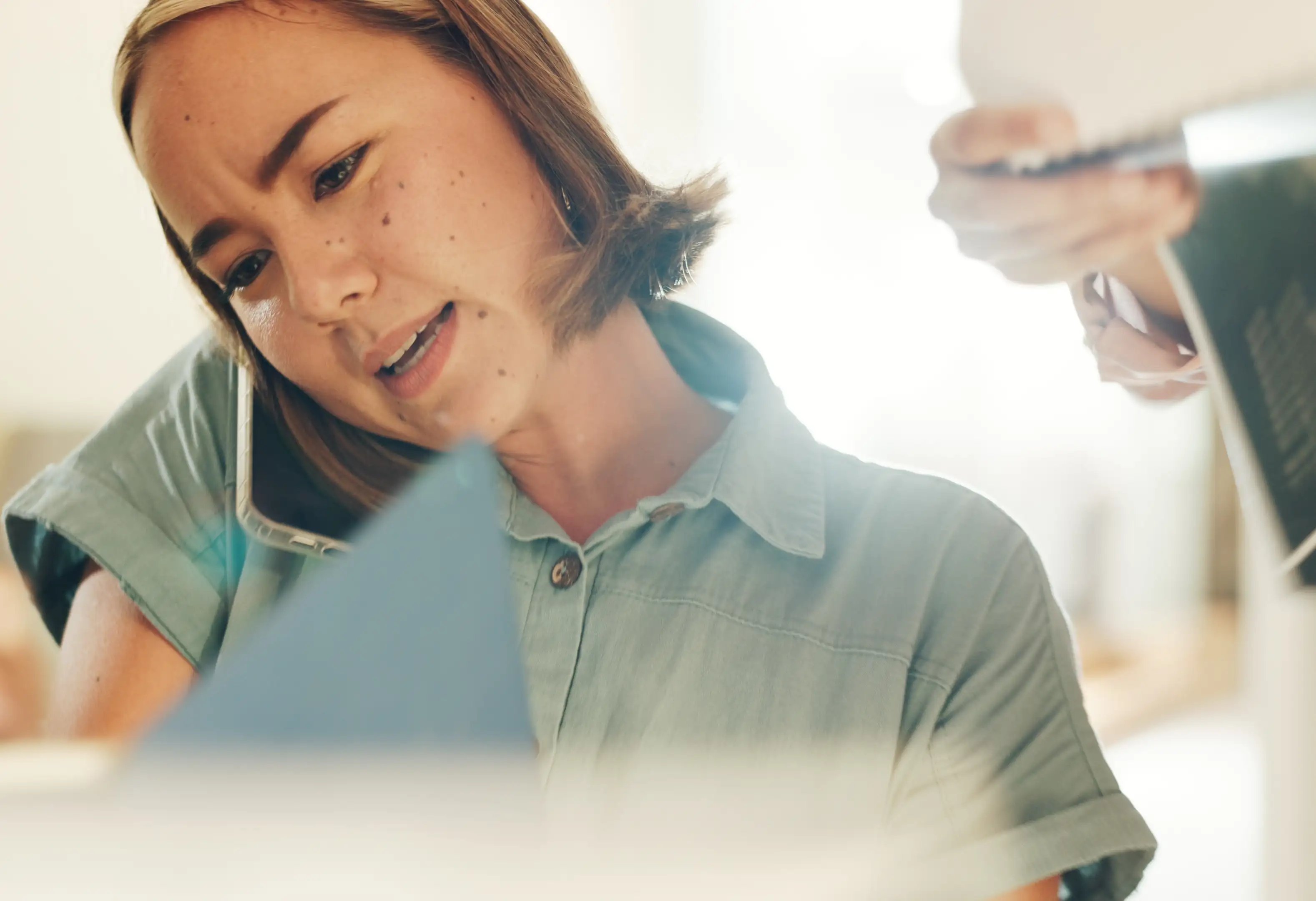 A stressed woman on the phone, while looking at a document, as another person waits with a laptop