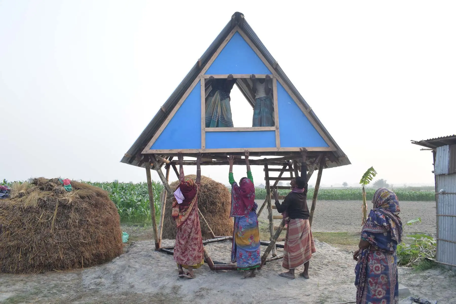 A group of Bangladeshi people dressed in colourful sarees assemble a bamboo house. Three people stand at the base with their arms stretched overhead, attaching a panel to the bamboo frame, while two people inside the structure help hold the panel up.