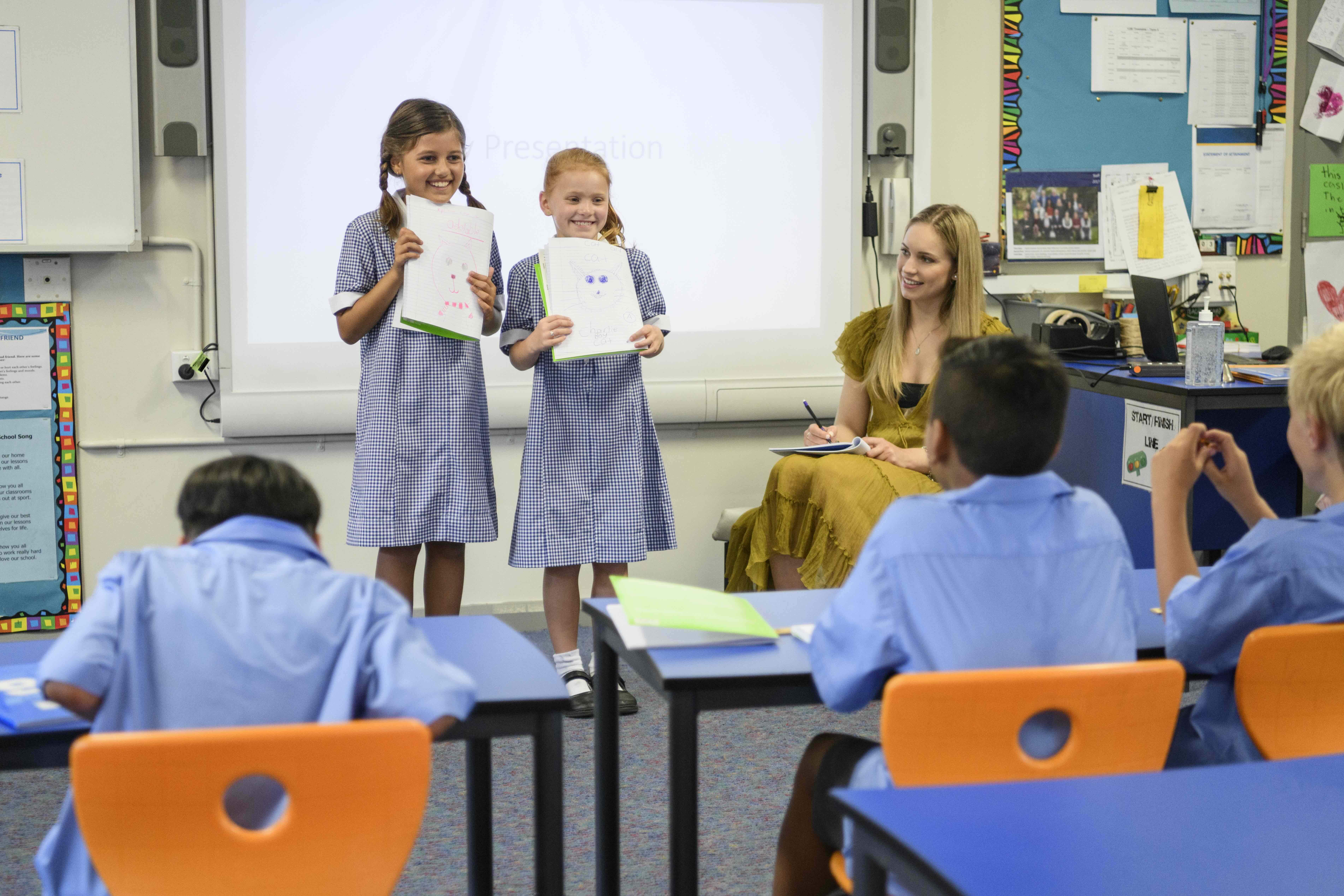 Two school girls presenting their work to the class with their teacher watching 