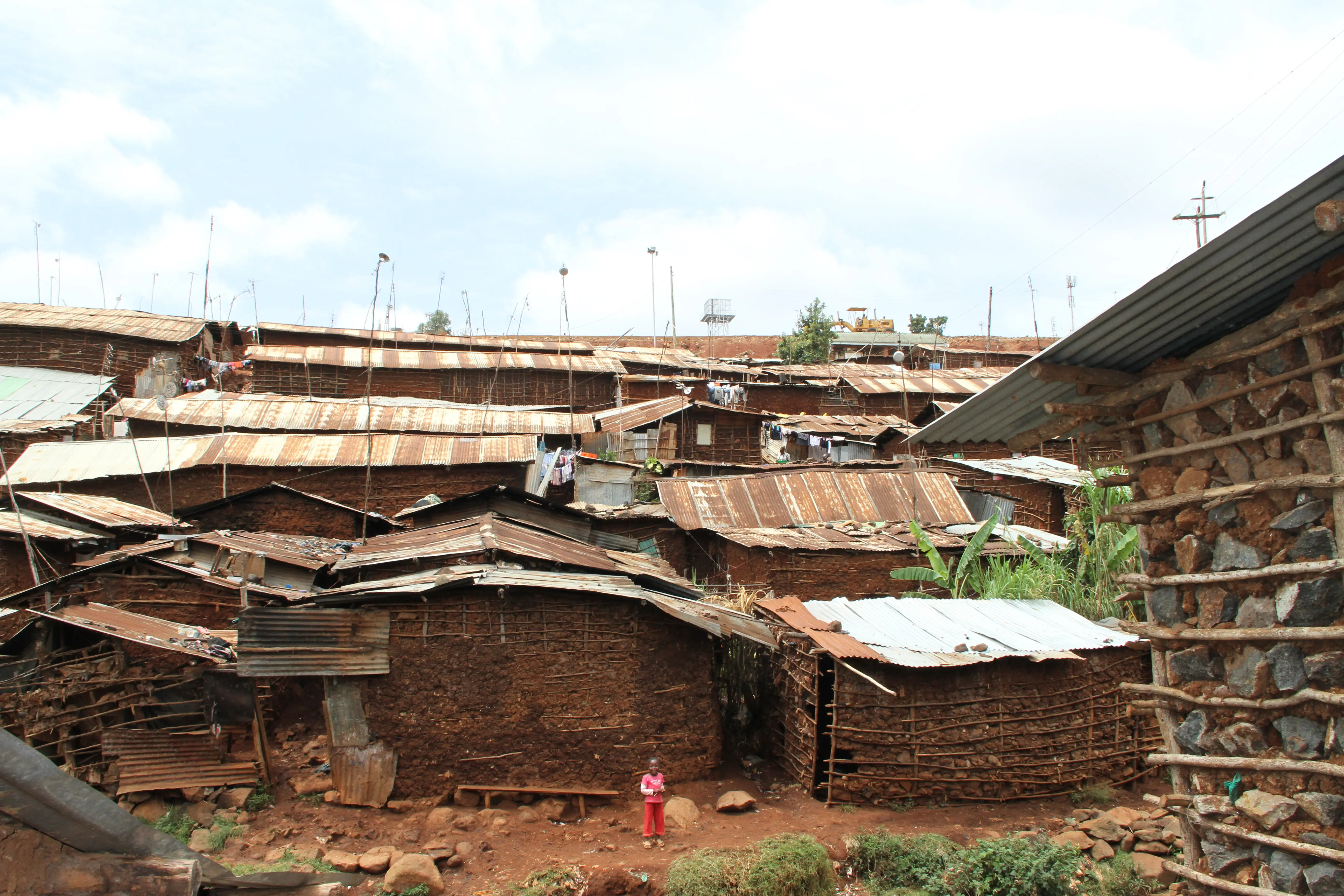 The brown roofs and stone walls of many buildings in the Kibera, Nairobi stretching towards the horizon. A girl in a red outfit stands in the distance smiling towards the camera.