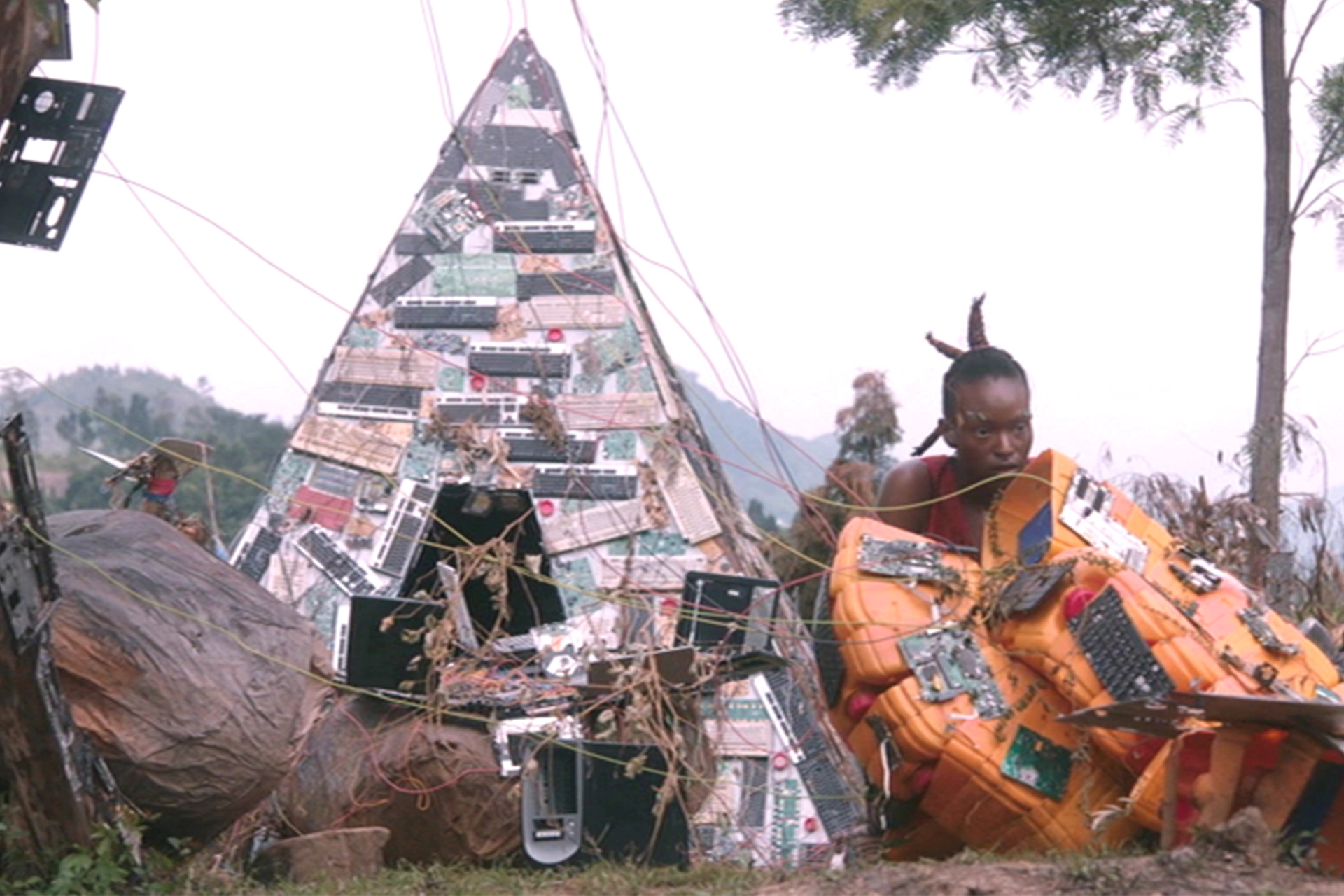 Still from Neptune Frost film, showing an African woman sitting in an outcast village. A makeshift shelter in the background is made of old electronic waste like keyboards