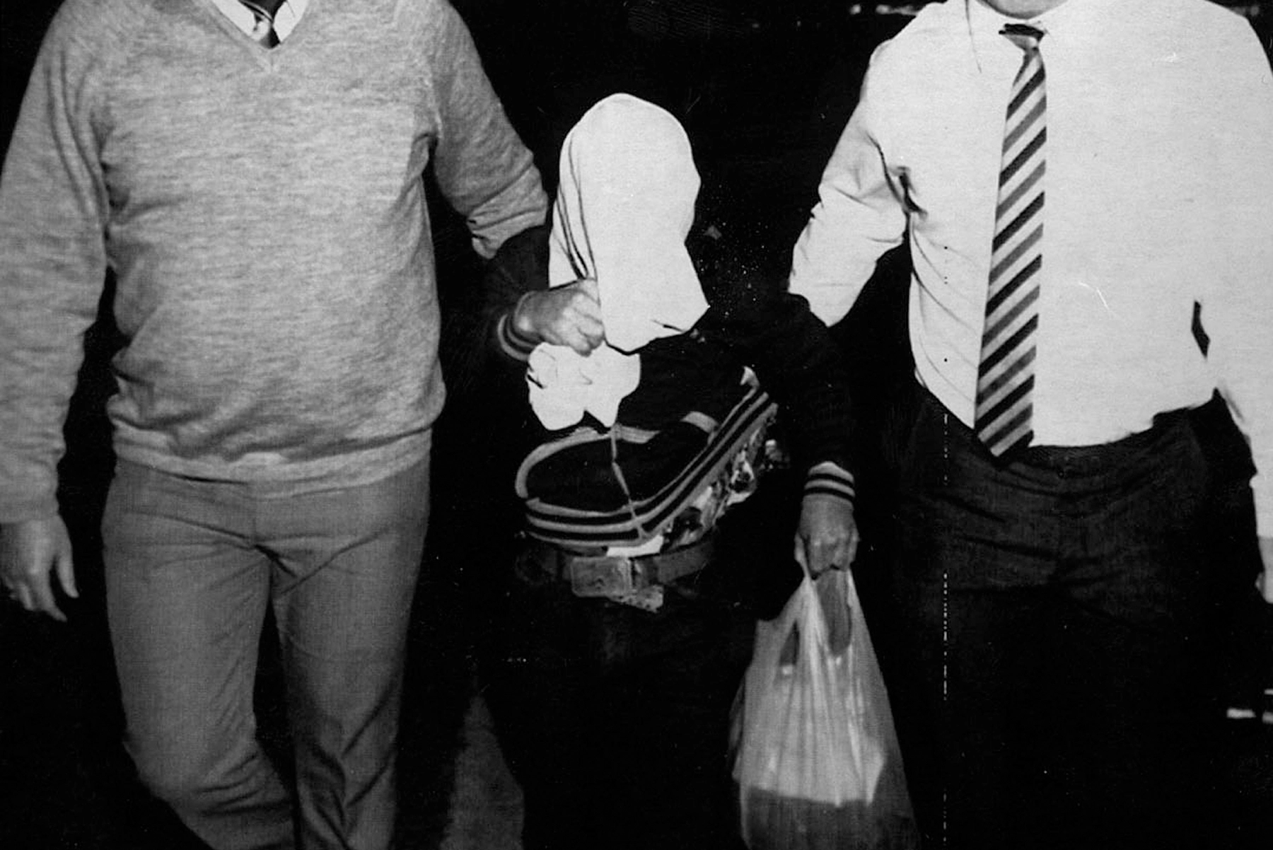 A man with a cloth over his head is arrested by two men, whose faces are cut off from the image