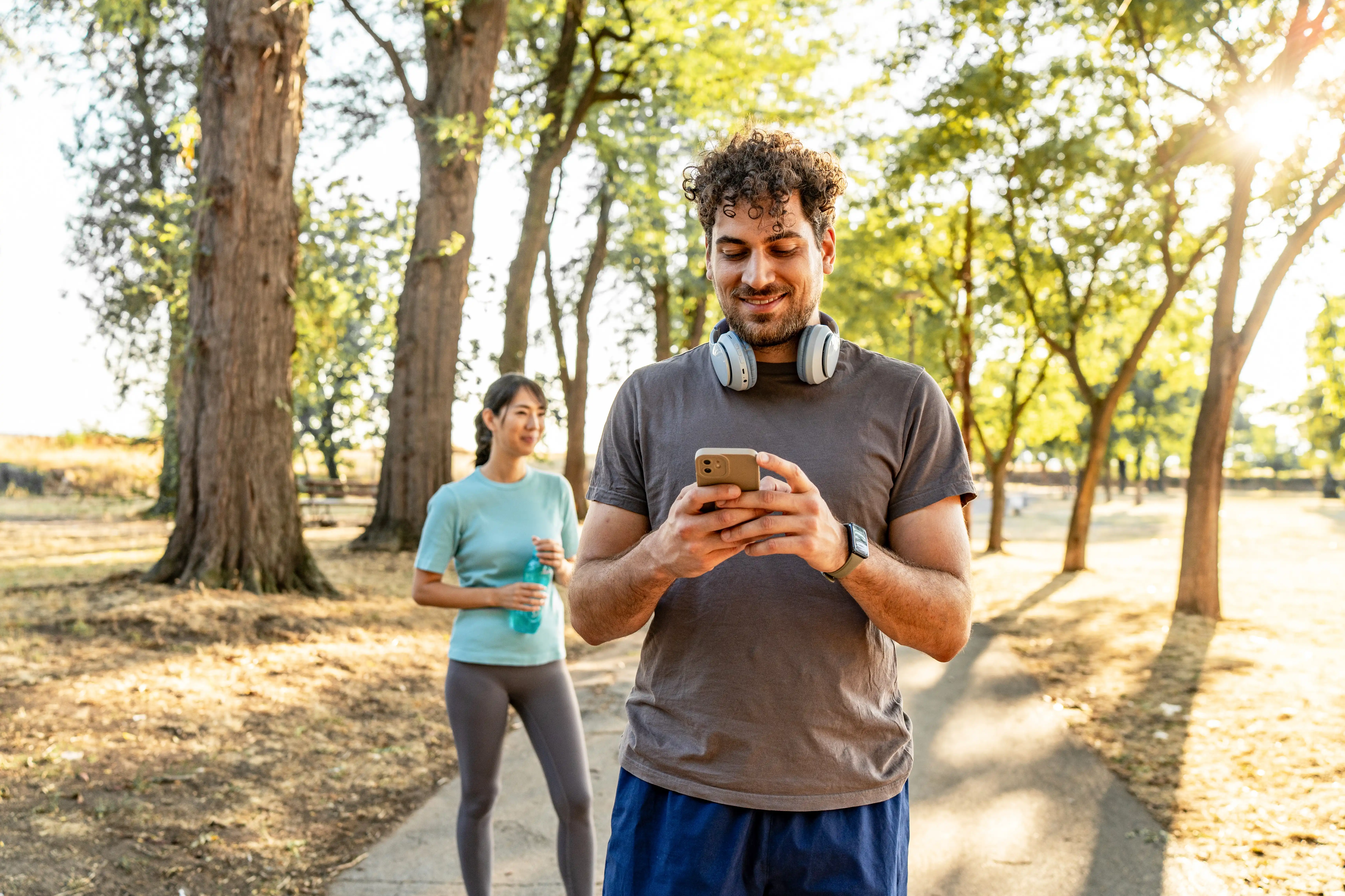 A man stands on a path out in nature in exercise clothes looking down and smiling at his phone