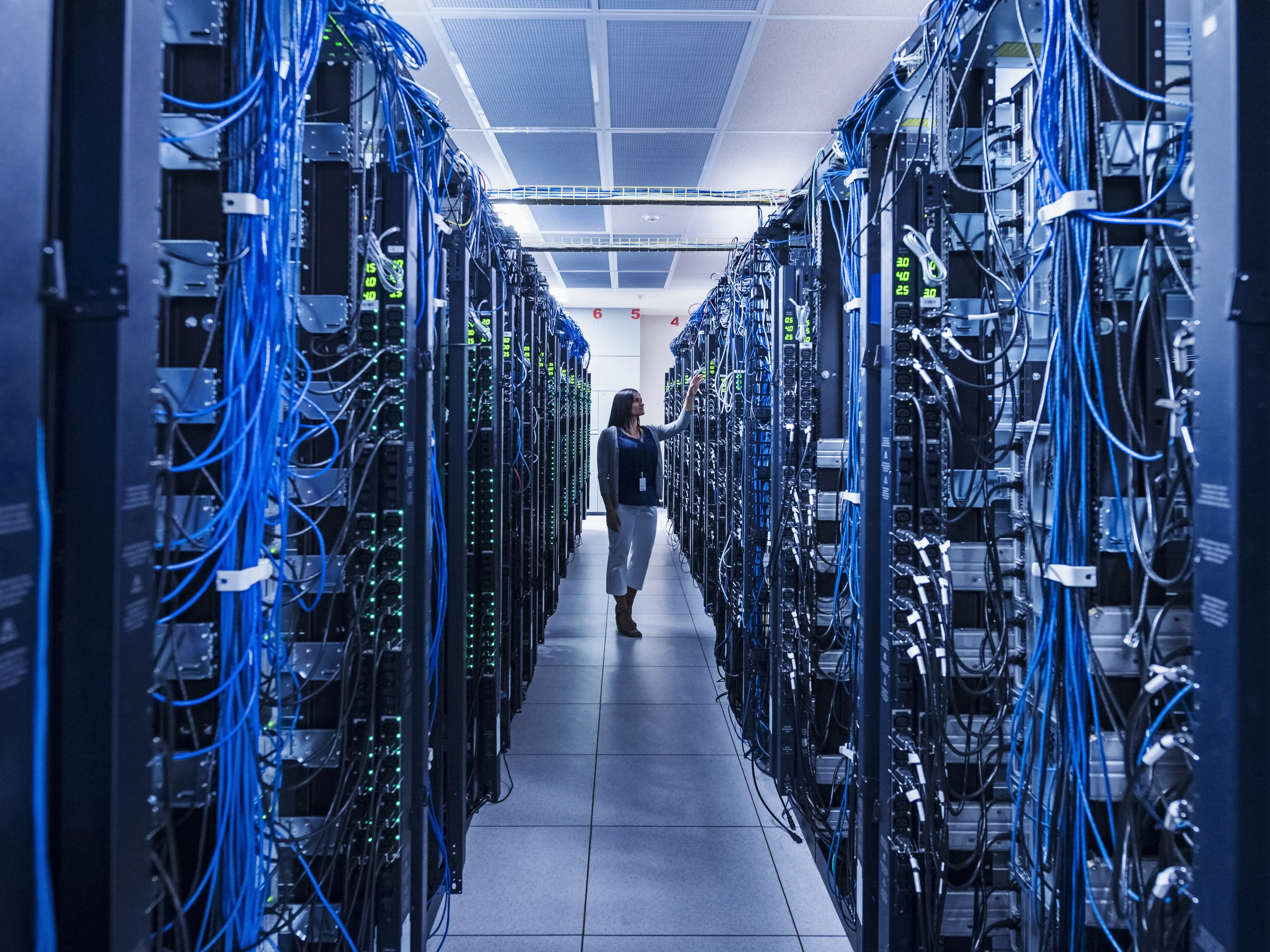 A woman stands in the aisle of a large server room, with blue cables dangling from the many machines