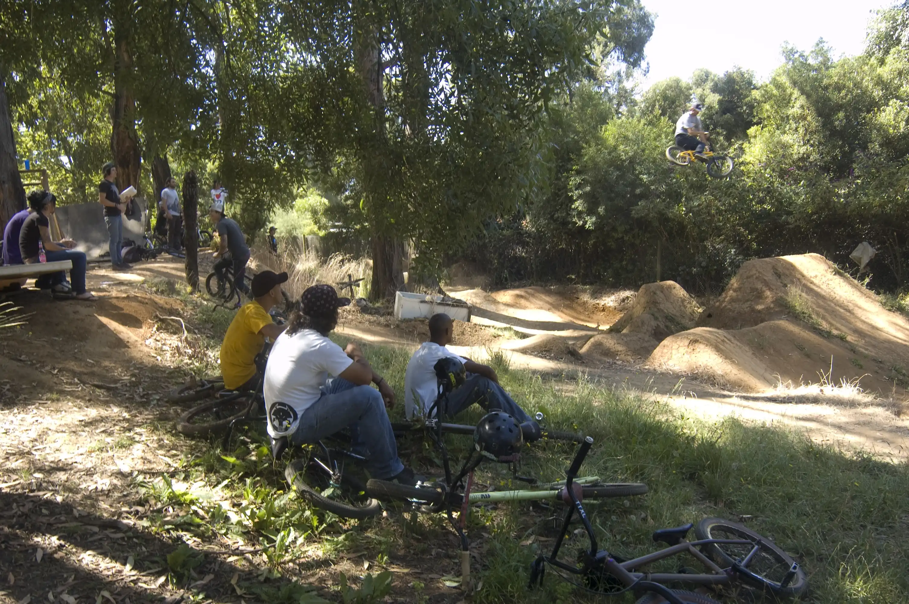 Three men sitting on a grassy hill with their bikes watch a friend who is high in the air coming off a jump at a BMX dirt track. There is bushland around and others watching the rider in the background.