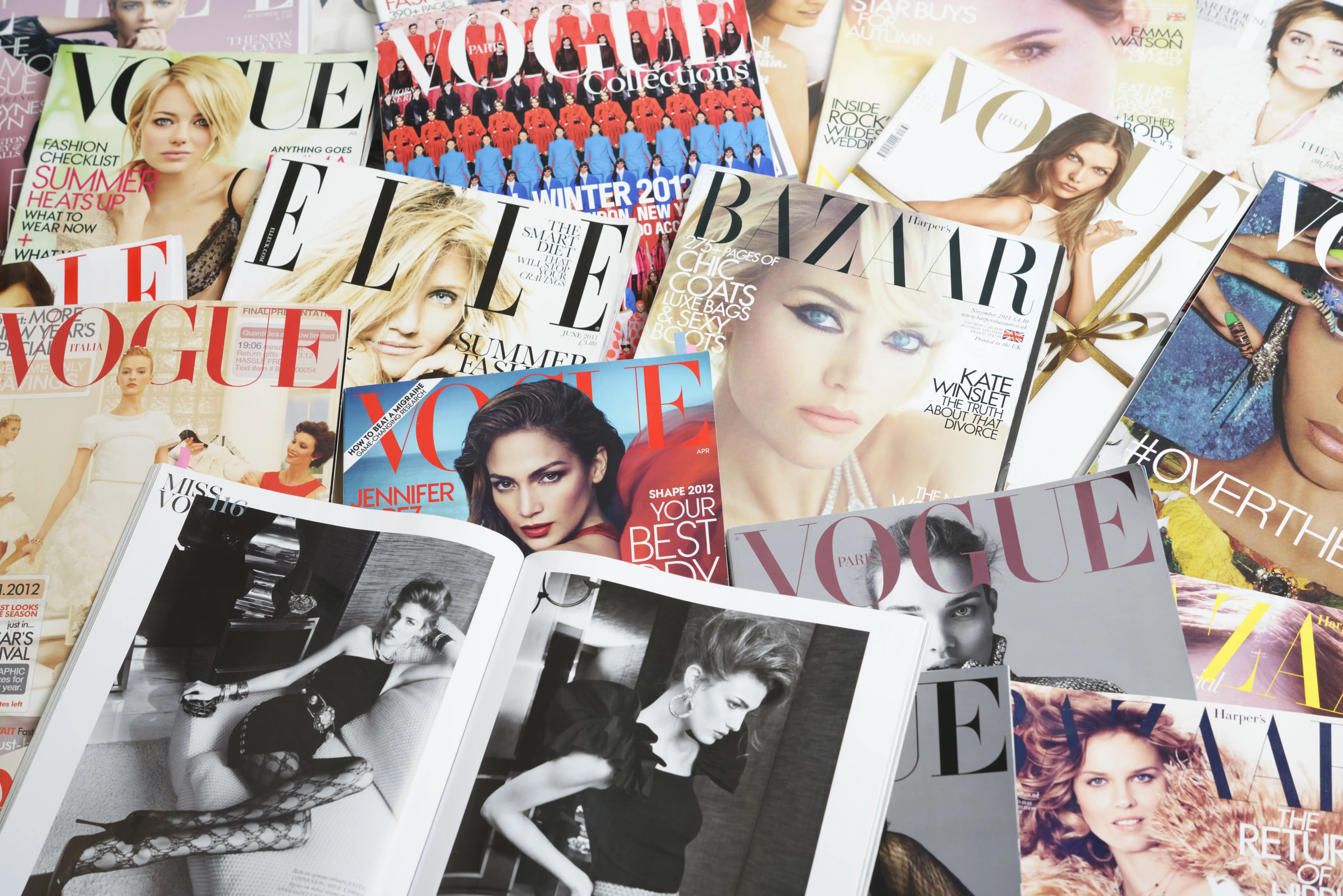 Various fashion magazines spread out across a table, showing front covers and inside others. Titles include Vogue, Elle and Bazaar.