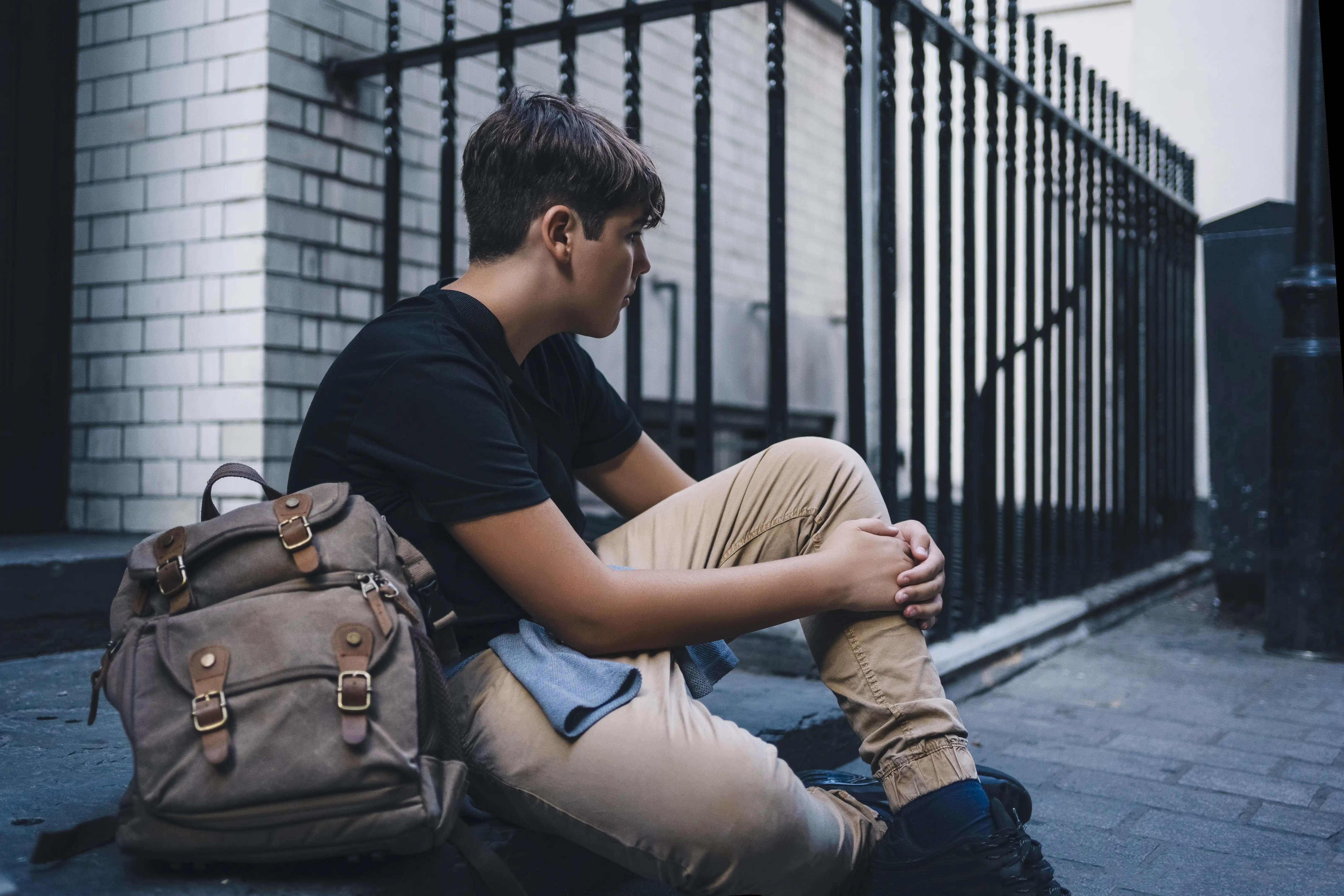 A teenager sitting on the street with a backpack looking sad