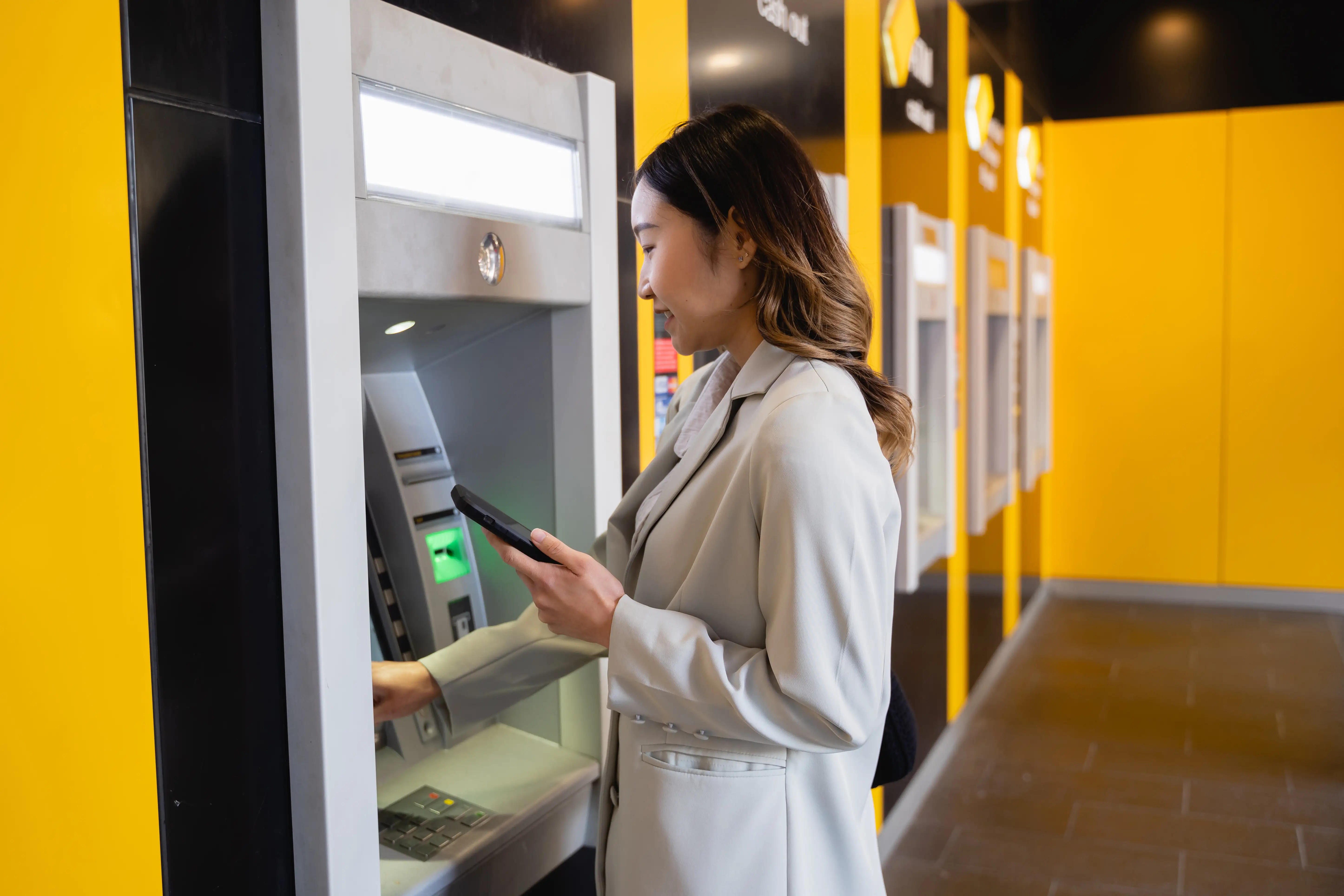 Young woman using her mobile phone while withdrawing money at an ATM machine 