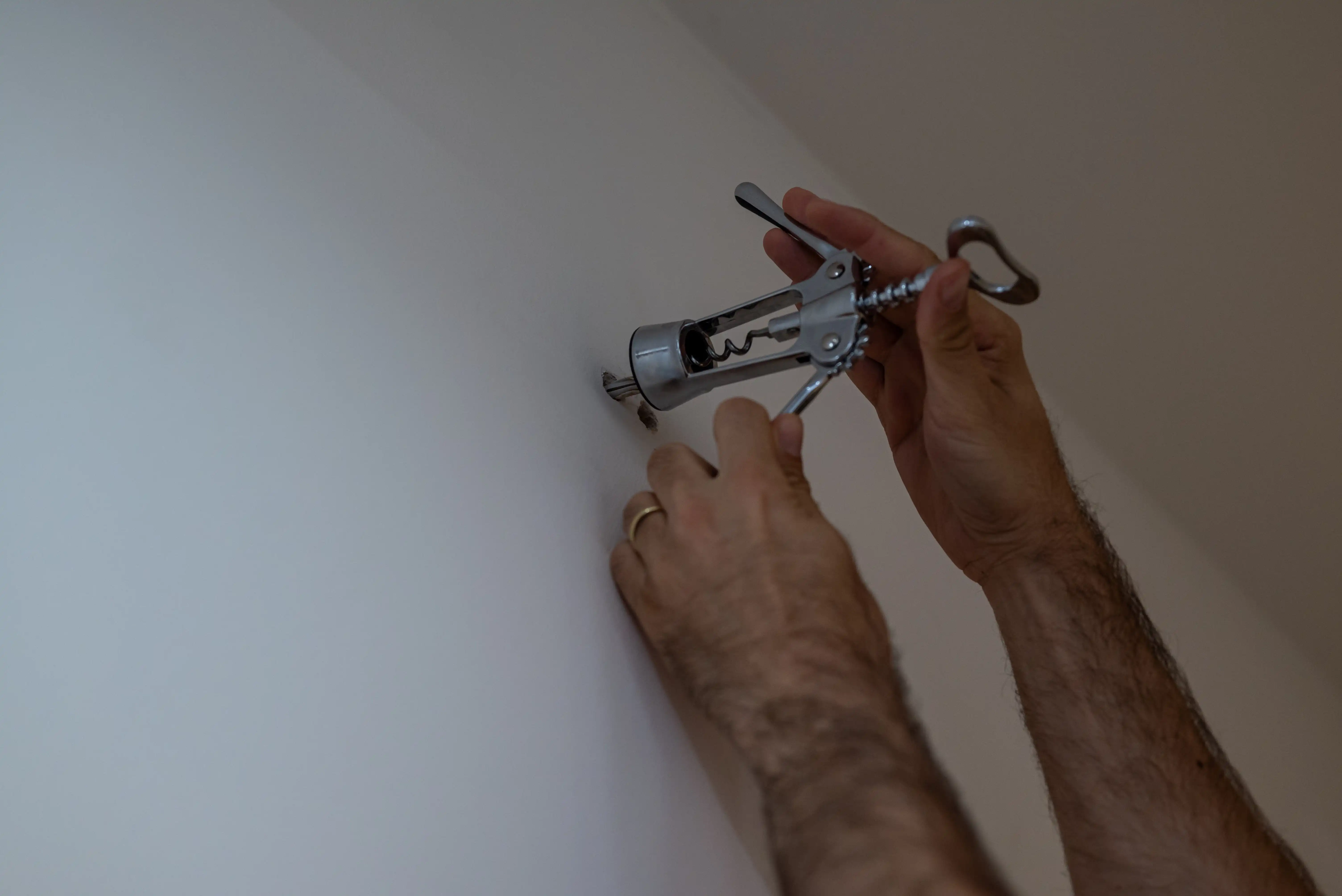 A man uses a corkscrew from a bottle opener to make a hole in his wall to hang a painting