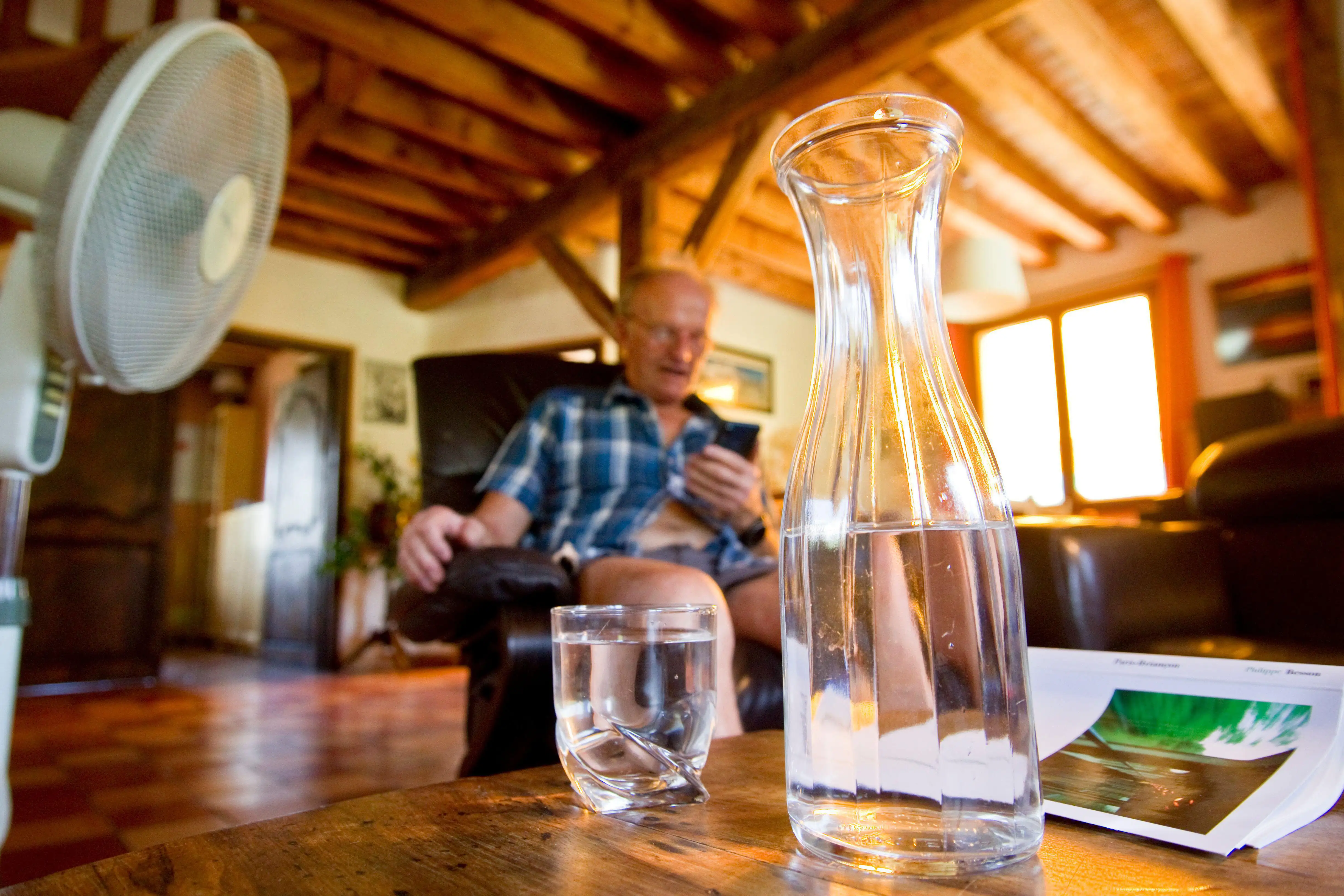 An elderly man sits in front of a fan with a bottle of water on the table