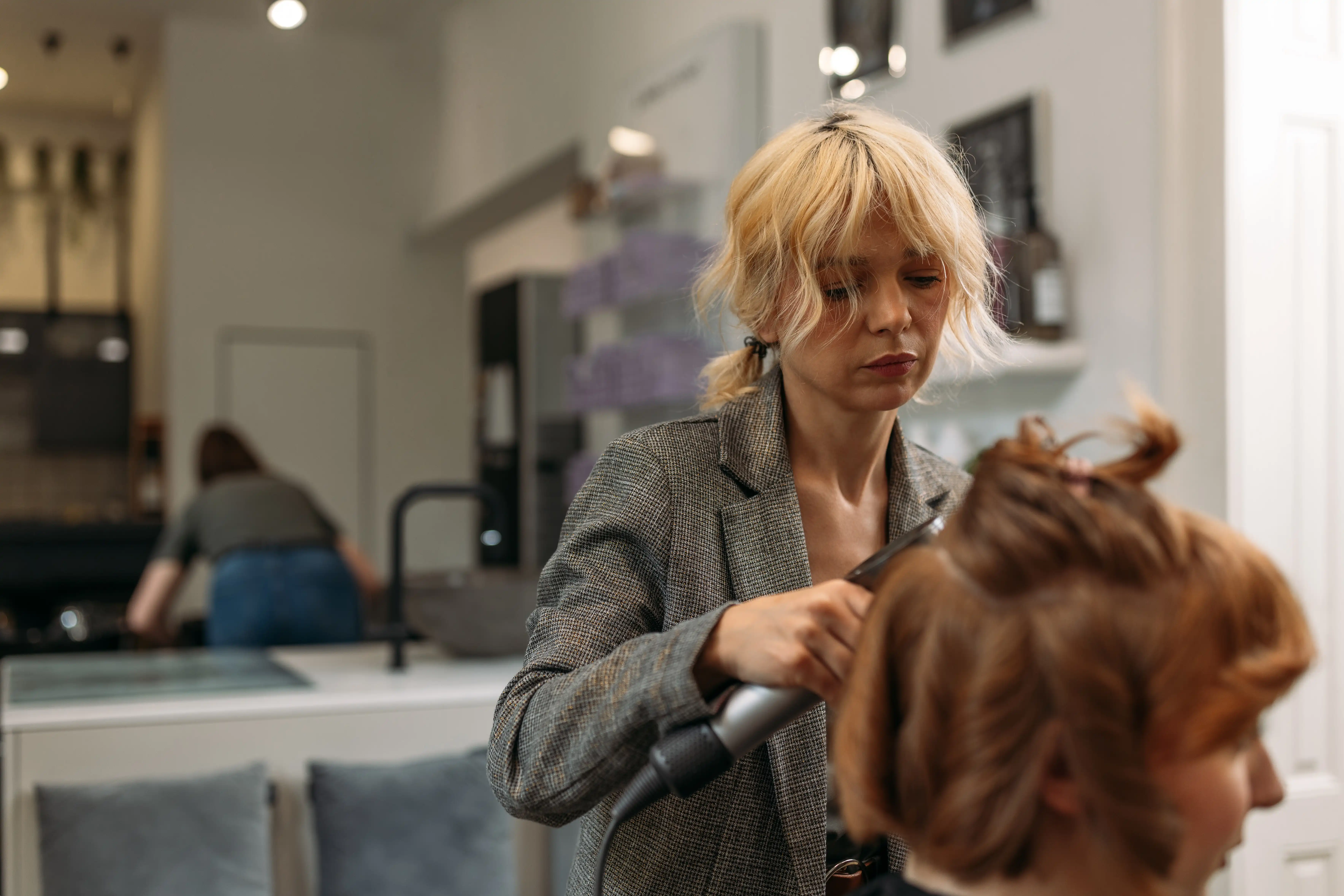 A female salon worker with frizzy hair in a blazer concentrating on curling a client’s hair