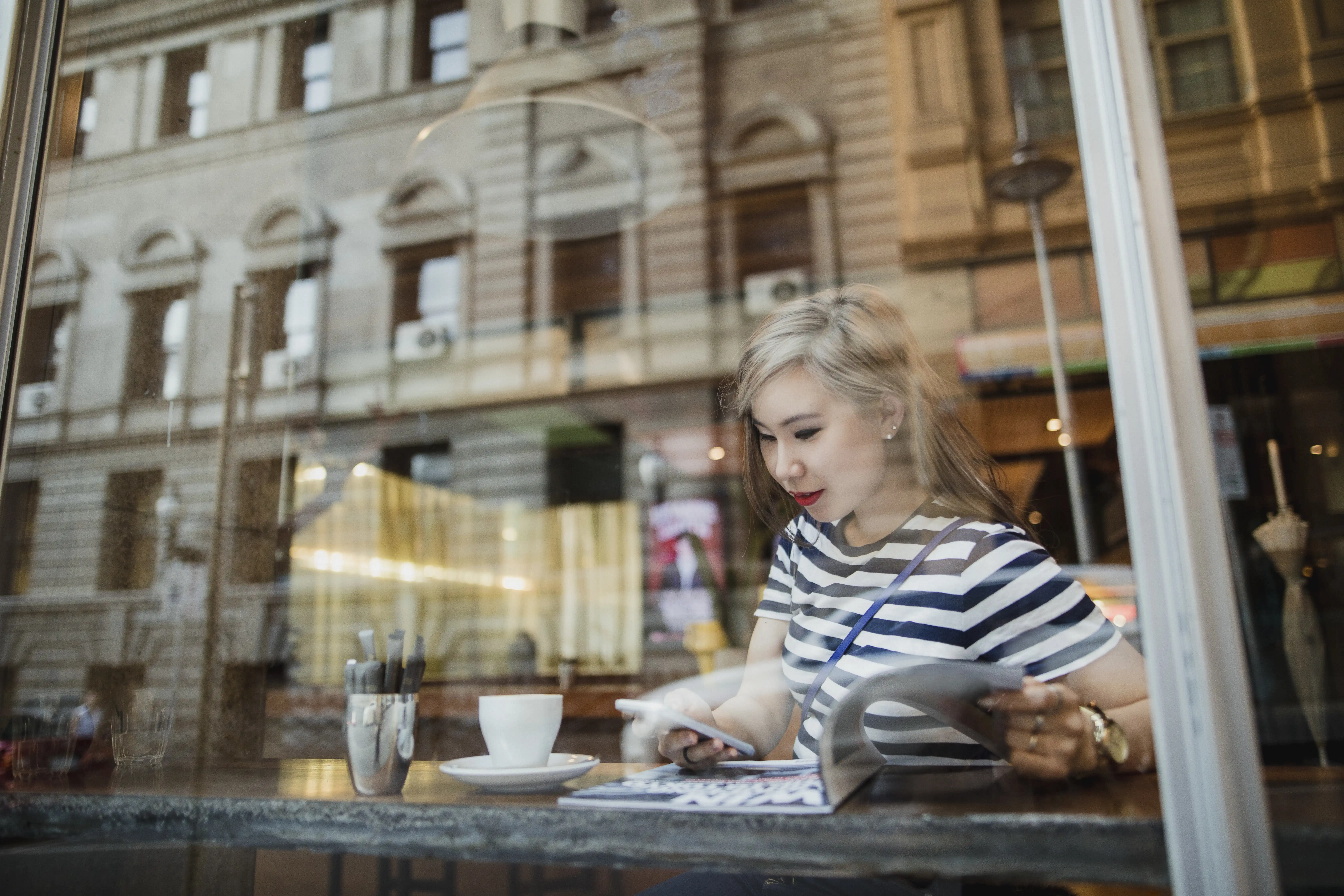 Young Asian woman sitting in a coffee shop reading a magazine and enjoying a cup of coffee. The building opposite is reflected in the glass the photo is taken through. 