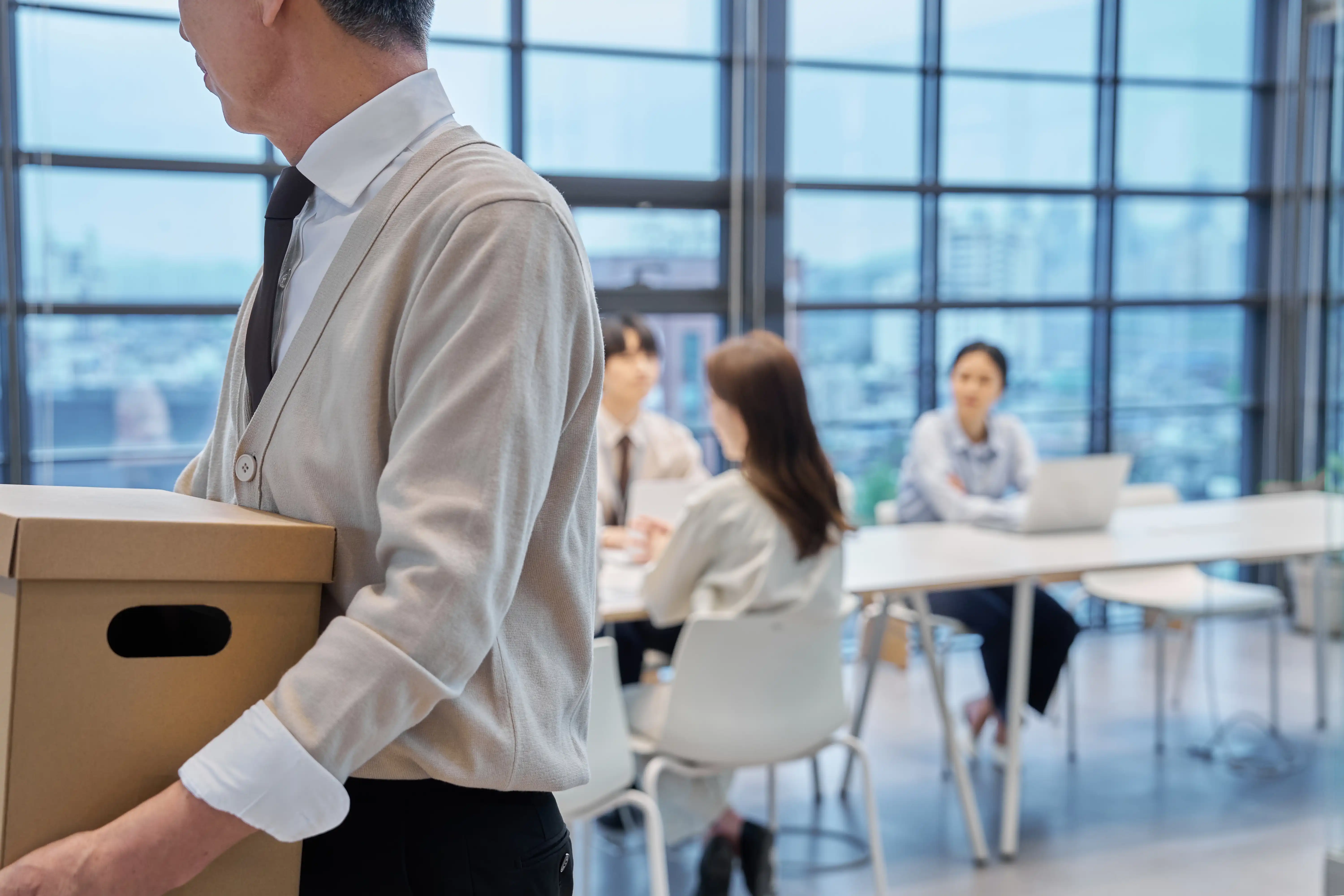 A man holds a cardboard box of his belongings as he looks back at an office room full of young people