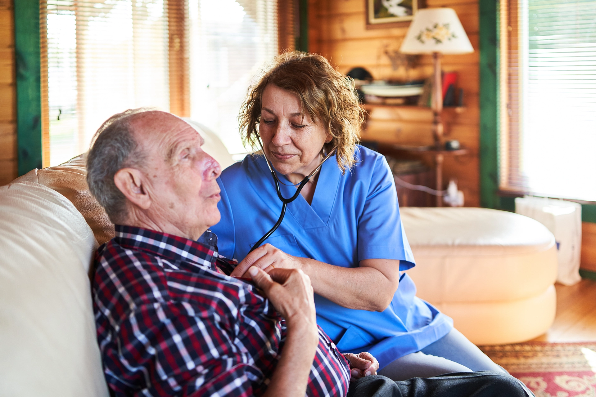 Middle aged nurse wearing scrubs checks elderly man's heart in his home using stethoscope