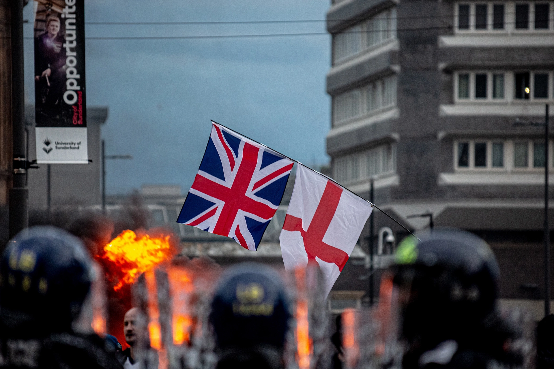  Riot scene in the UK, showing riot police wearing helmets, British and English flags being waved, and fire explosion in the background