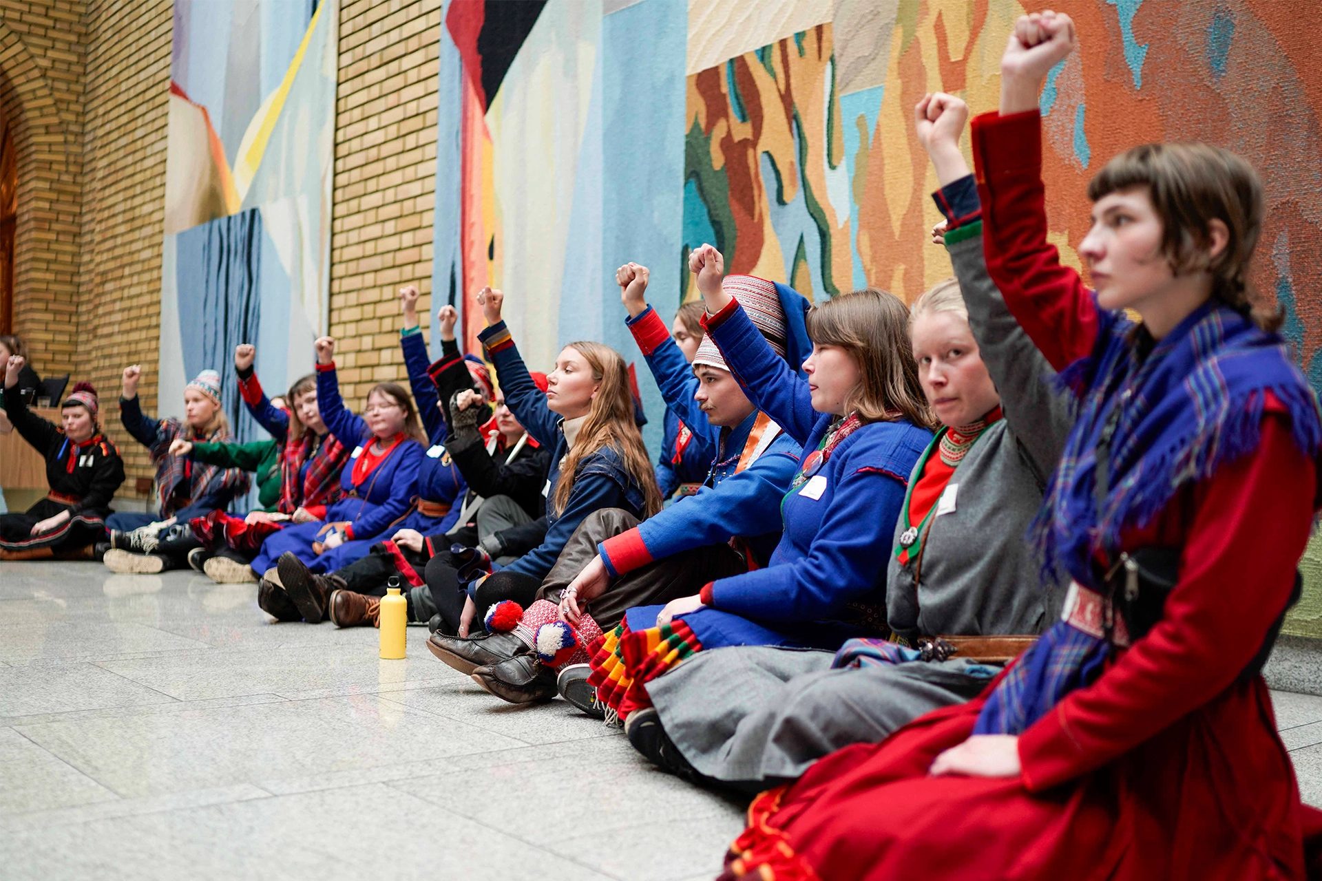 Group of women in traditional Sami dress, sitting on floor of Parliament in Norway with their fists raised in the air