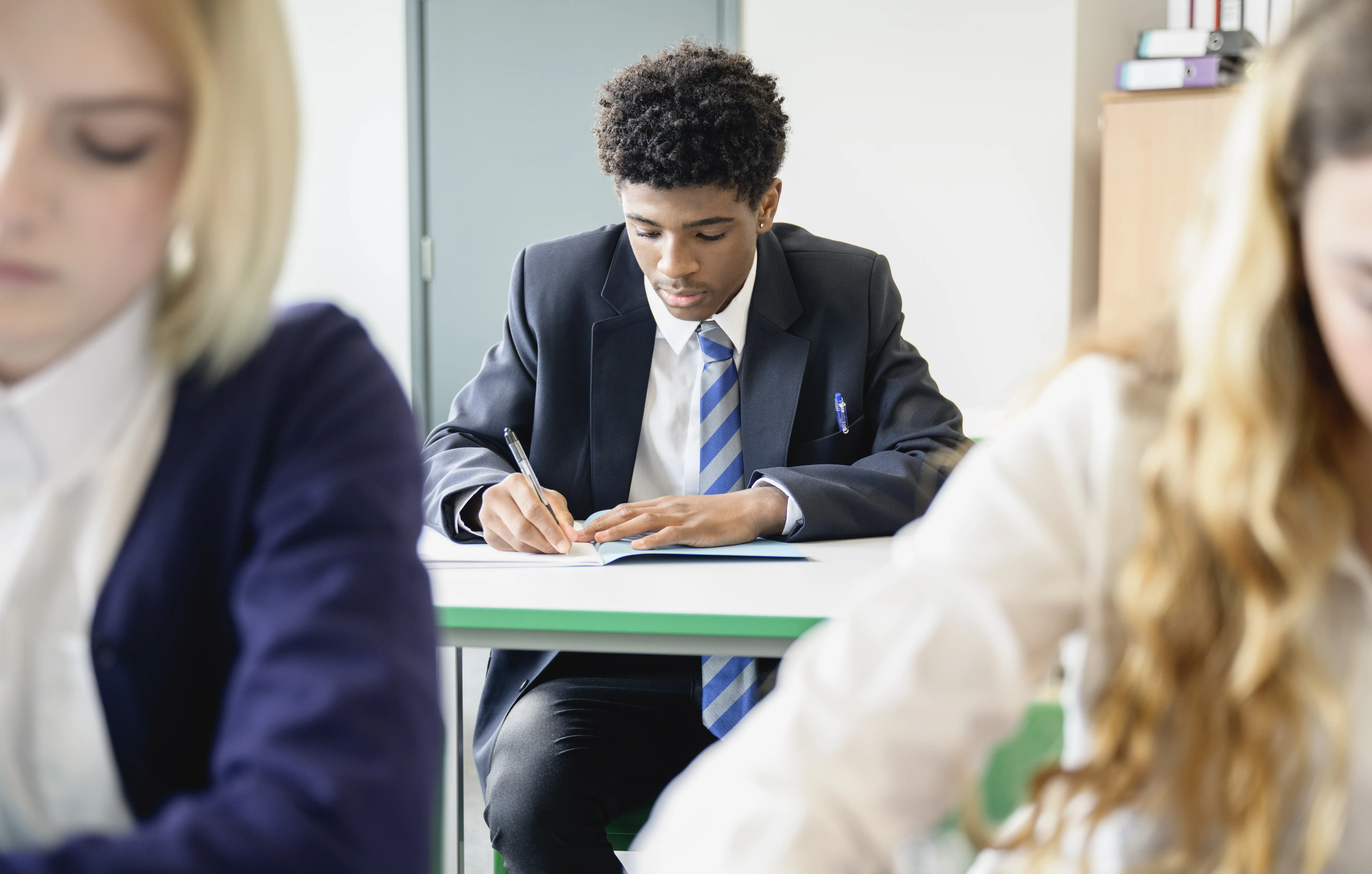 A teen boy in uniform sitting at secondary school desk and concentrating while writing