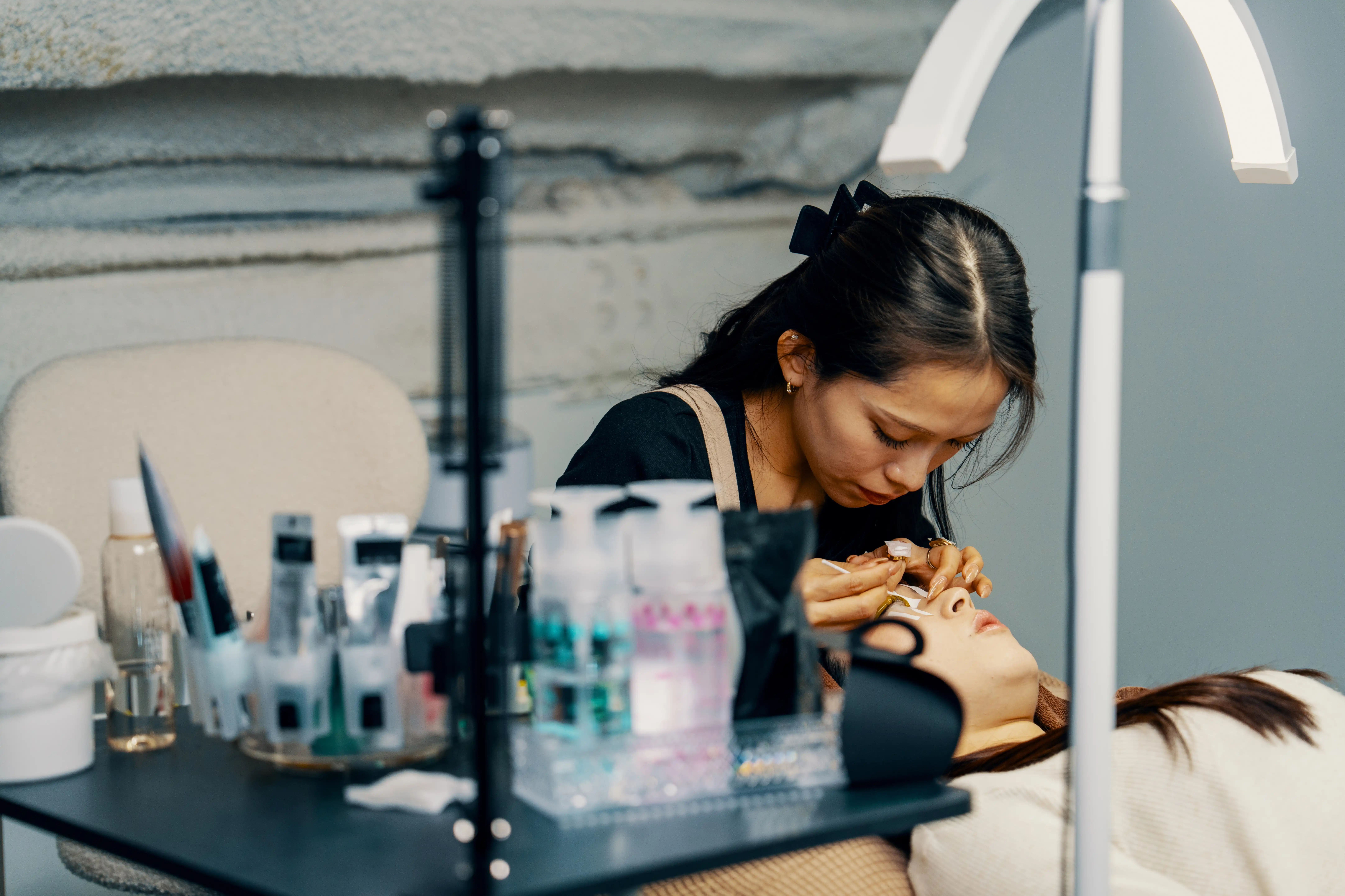A beautician leans over a client who is laying down as she puts fake eyelashes on her. A table of beauty products sit beside them