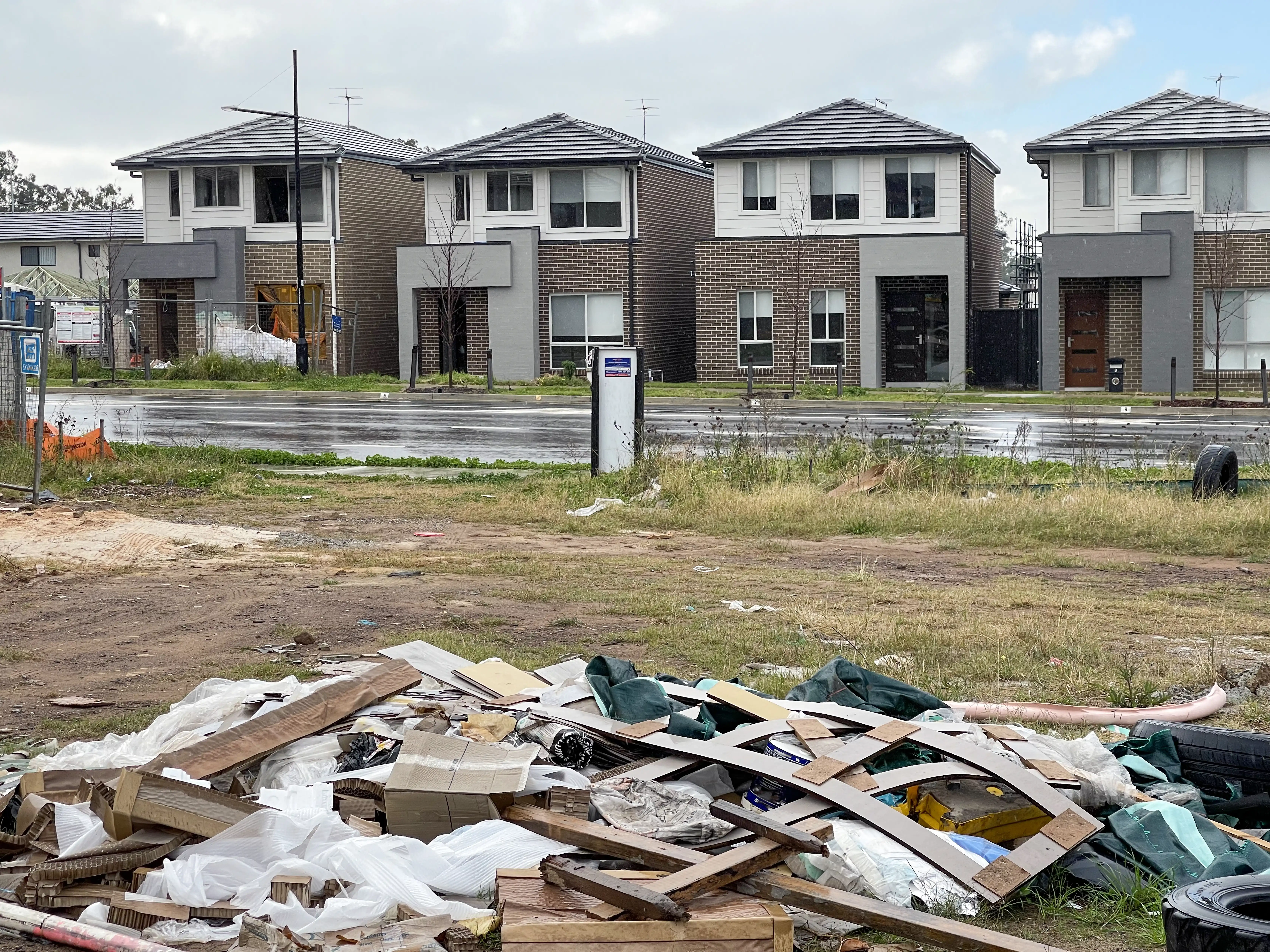 An empty construction site full of timber and plastic waste, looking over the road to a new housing development. Each of the houses look similar and are made from brick and concrete.