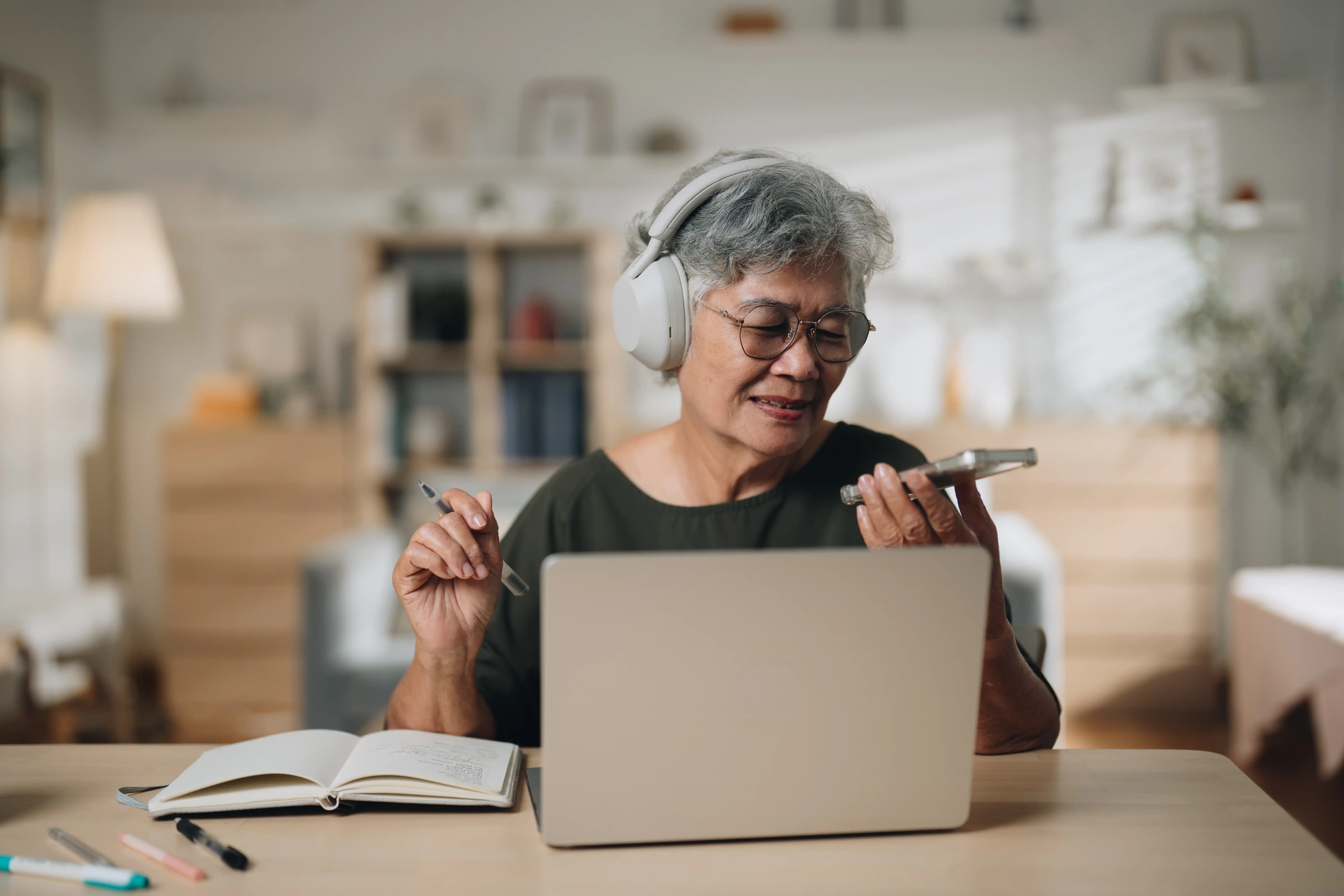 A senior woman holding her phone to her mouth while she wears headphones and sits in front of a laptop, using technology to help her work